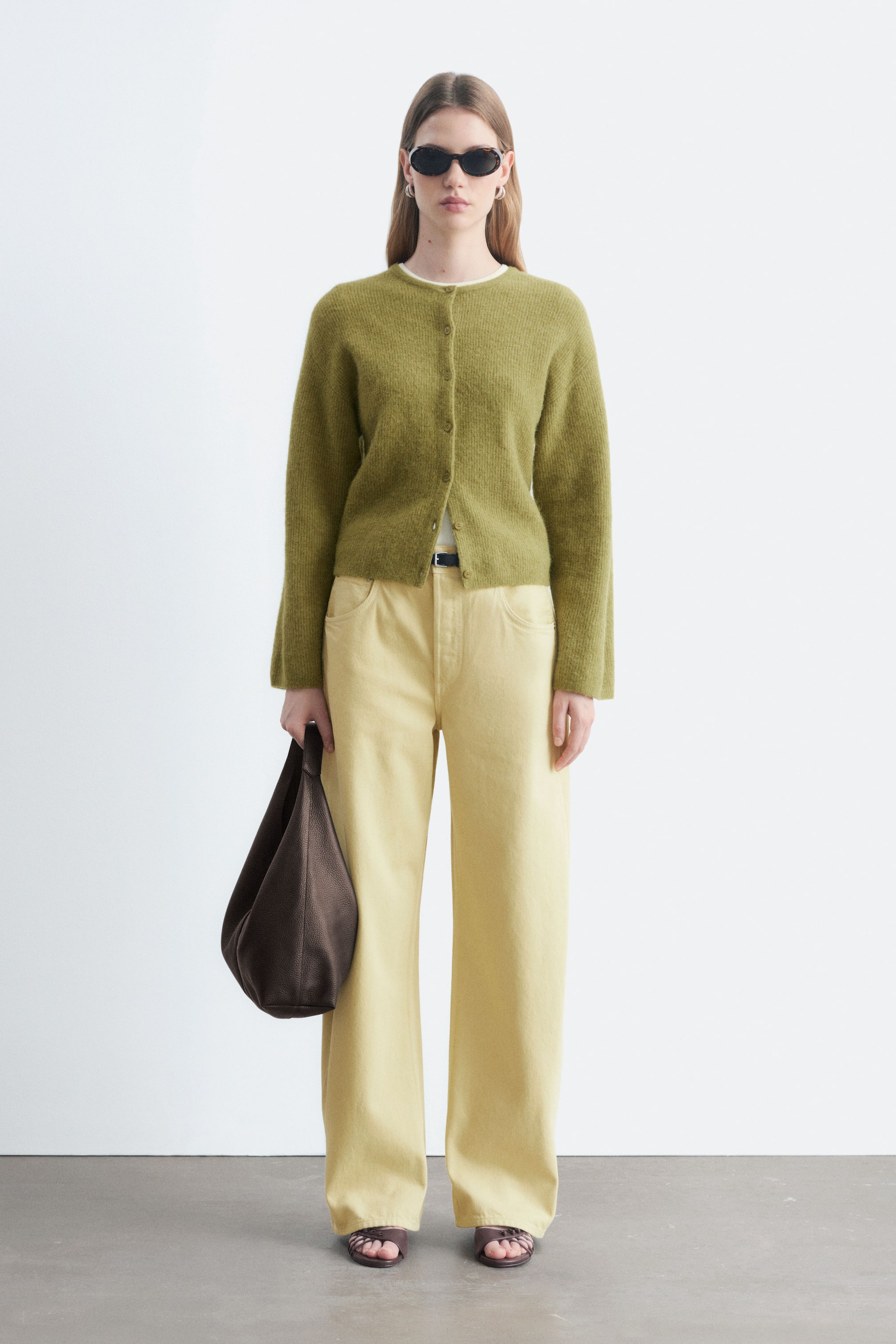 Visualizza l'immagine ingrandita: Una cardigan in maglia verde muschio con chiusura frontale a bottoni e girocollo è indossata dalla modella. È abbinata a pantaloni palazzo giallo pallido e a una borsa a mano in pelle marrone scuro portata in una mano.