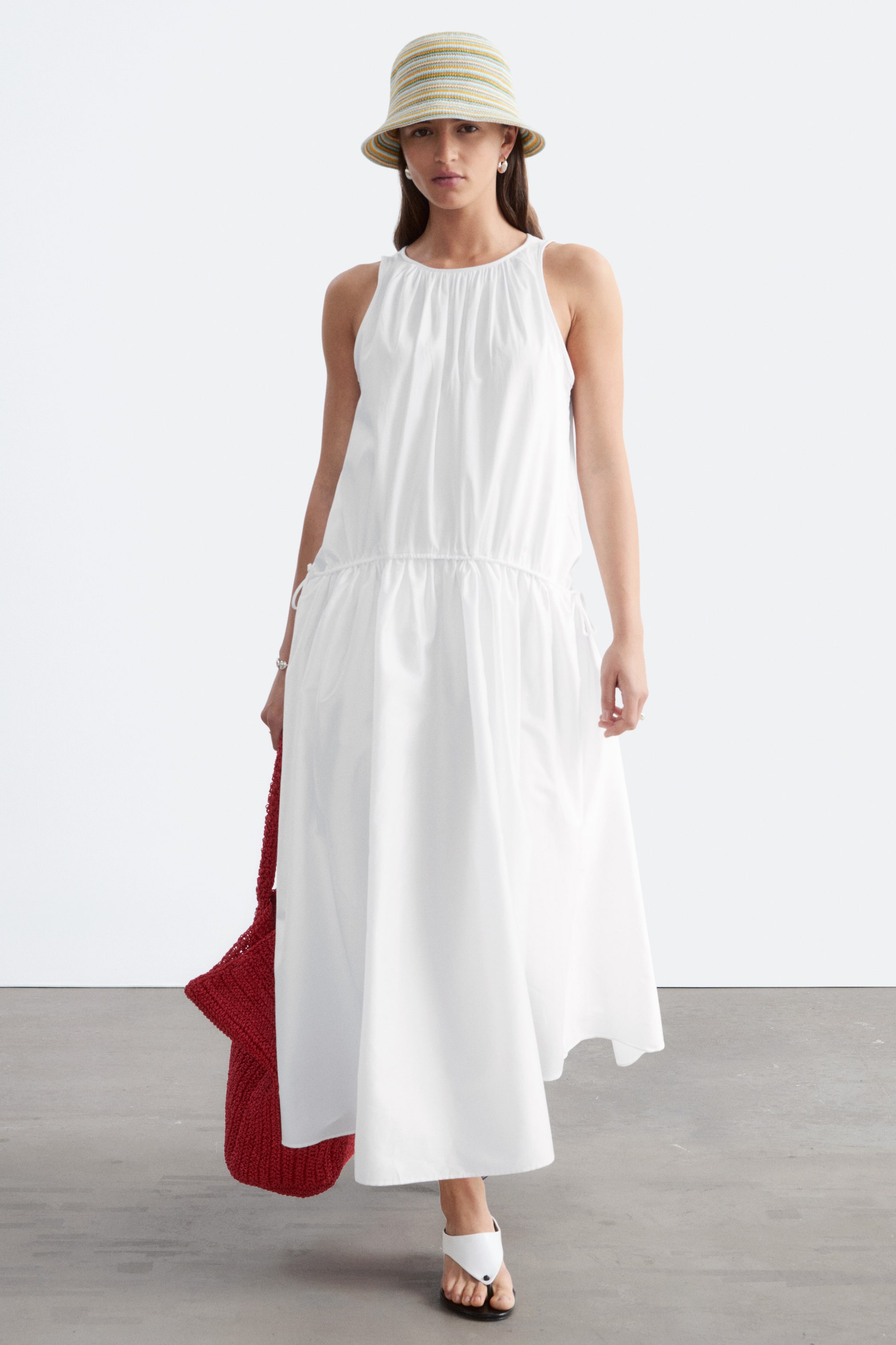 Ver imagen más grande: Un vestido blanco sin mangas, con frunces en el escote y un canal de cordón en la cintura con lazos, es llevado por la modelo. El vestido cae por encima del tobillo, creando una silueta fluida, y la modelo también lleva un sombrero de cubo a rayas y un bolso de ganchillo rojo.