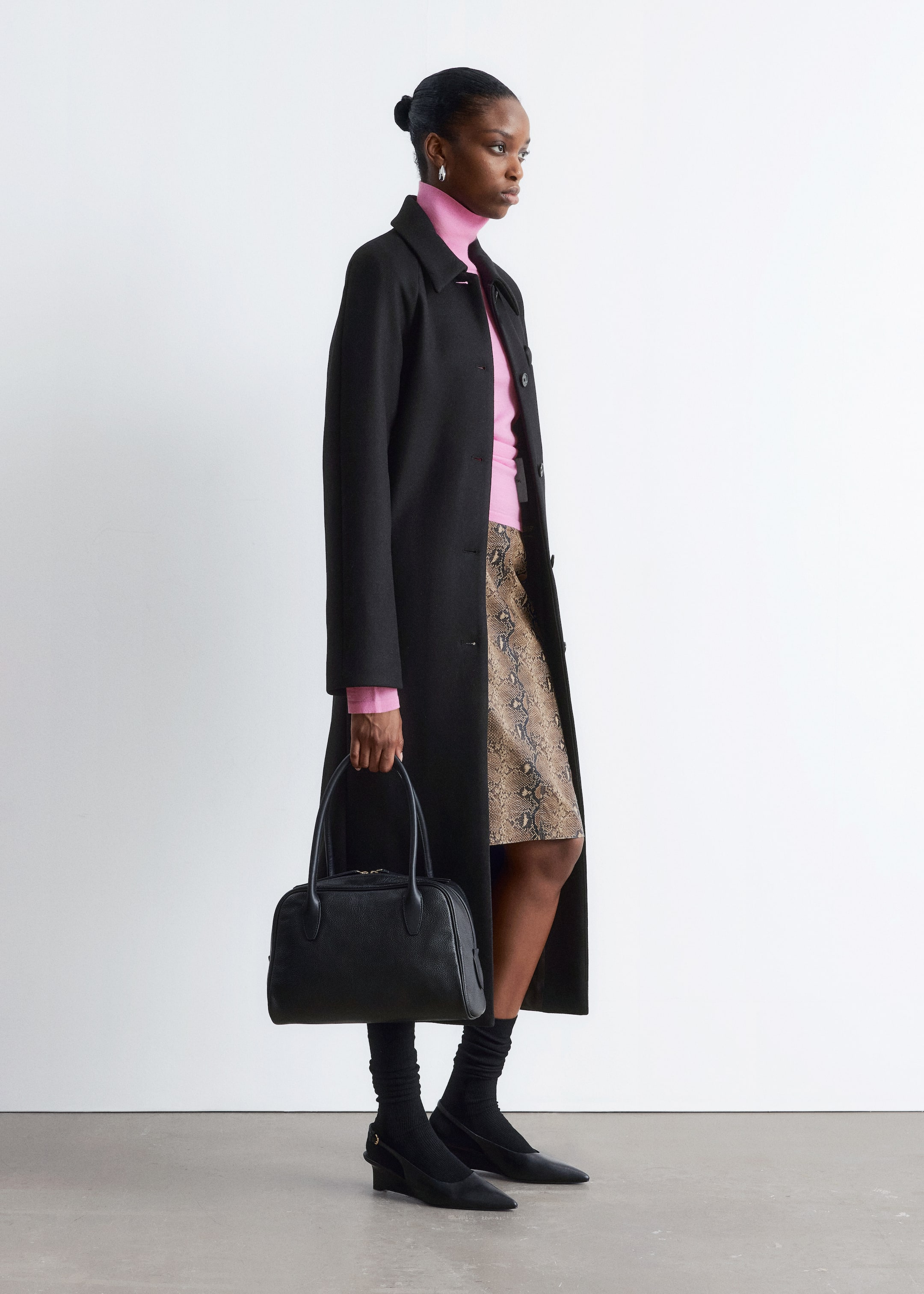 Visualizza immagine più grande: Un lungo cappotto nero monopetto con un colletto classico viene indossato, presentato di profilo per evidenziare la sua lunghezza e stratificazione su un maglione a collo alto rosa acceso e una gonna al ginocchio con un motivo a pelle di serpente marrone e nero. L'ensemble è completato da una borsa strutturata nera con due manici superiori e scarpe slingback a punta nera con zeppa.