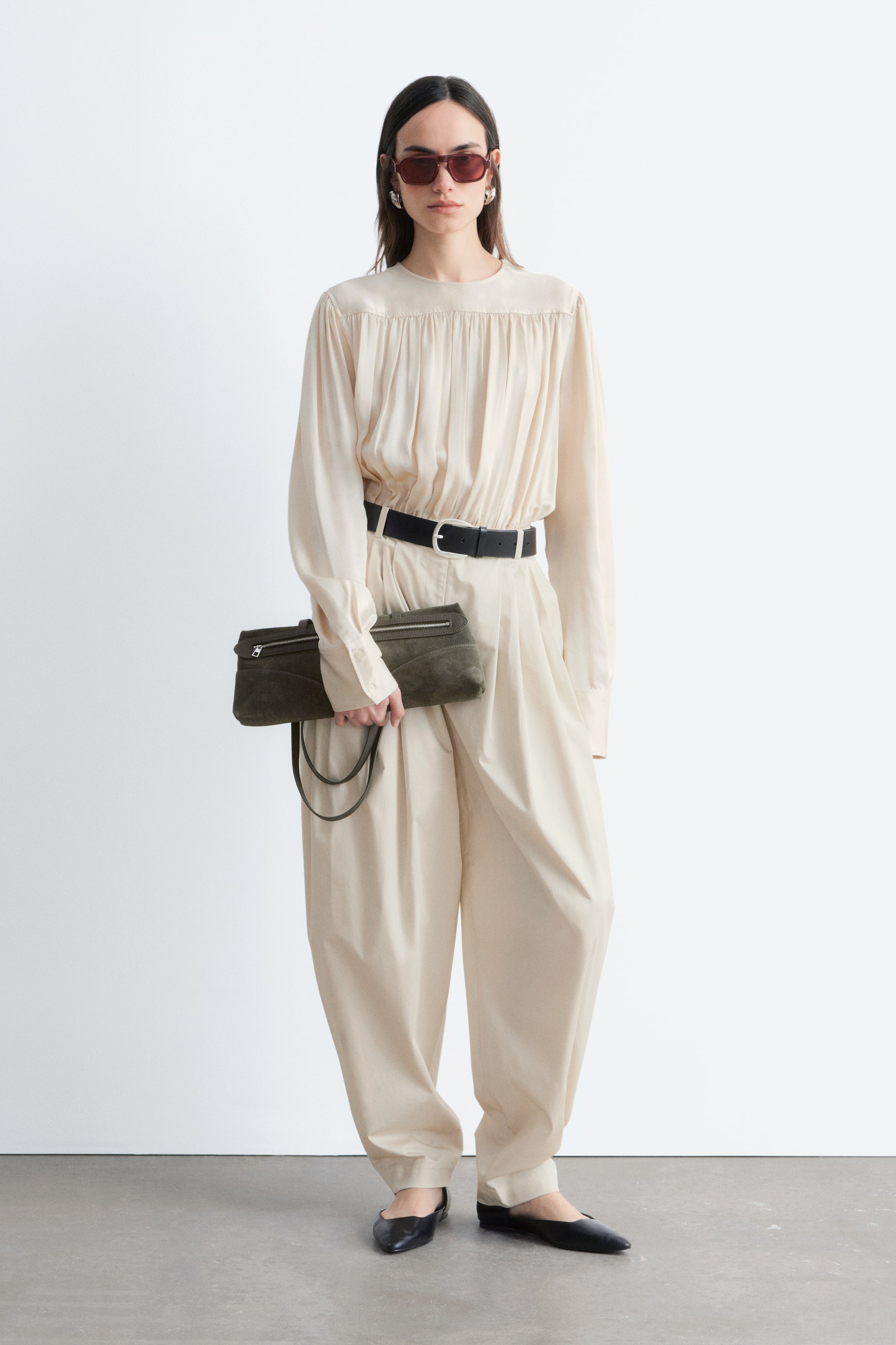 Se større billede: Modellen bærer højtaljede plisserede bukser i en lys beige farve. Bukserne har et rent linning med bæltestropper, og deres voluminøse ben smalner blidt ind mod anklerne, med et sort bælte trukket gennem stropperne.