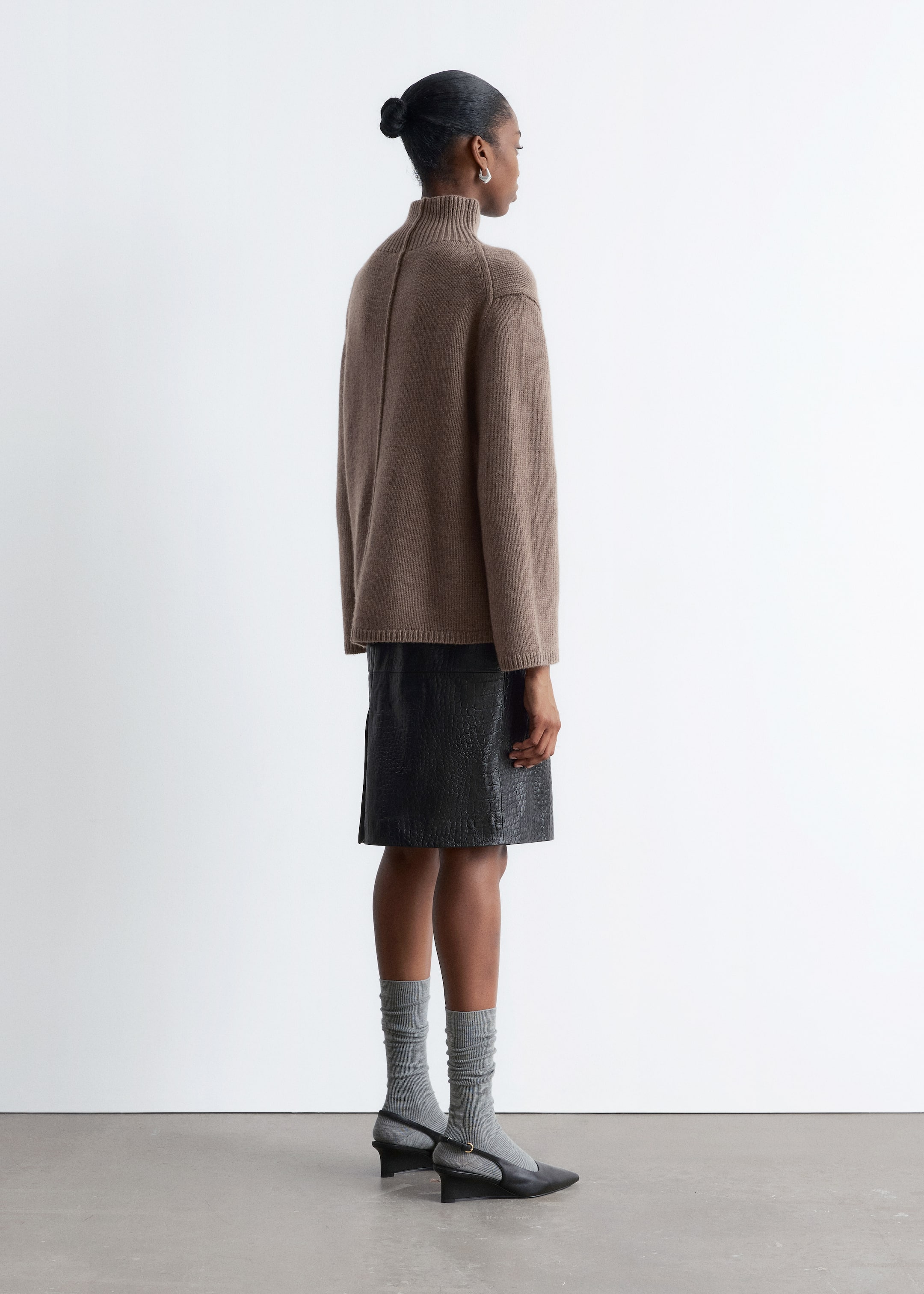 Grösseres Bild ansehen: Ein brauner Rollkragenpullover mit geripptem Kragen und Saum wird vom Model getragen. Der Pullover hat eine lockere, langärmelige Passform mit einer sichtbaren Naht, die den Rücken hinunterläuft, und wird über einem schwarzen, strukturierten Rock gestylt.