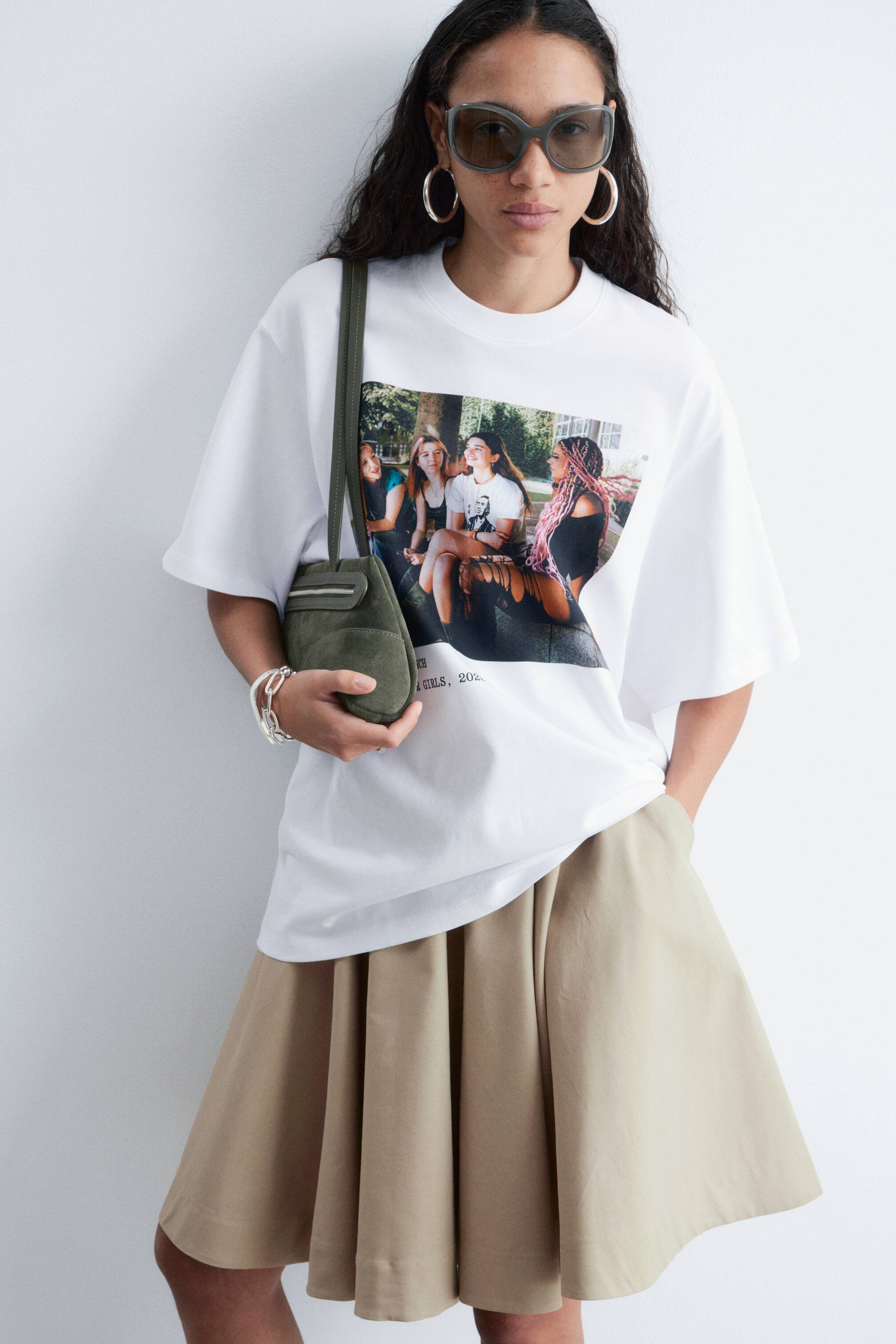 Se større billede: En hvid oversized T-shirt med et fotografisk tryk af fire personer og ordet "GIRLS" bæres af modellen. Modellen styler T-shirten med en plisseret khaki nederdel, og en lille grøn crossbody taske bæres over T-shirten.