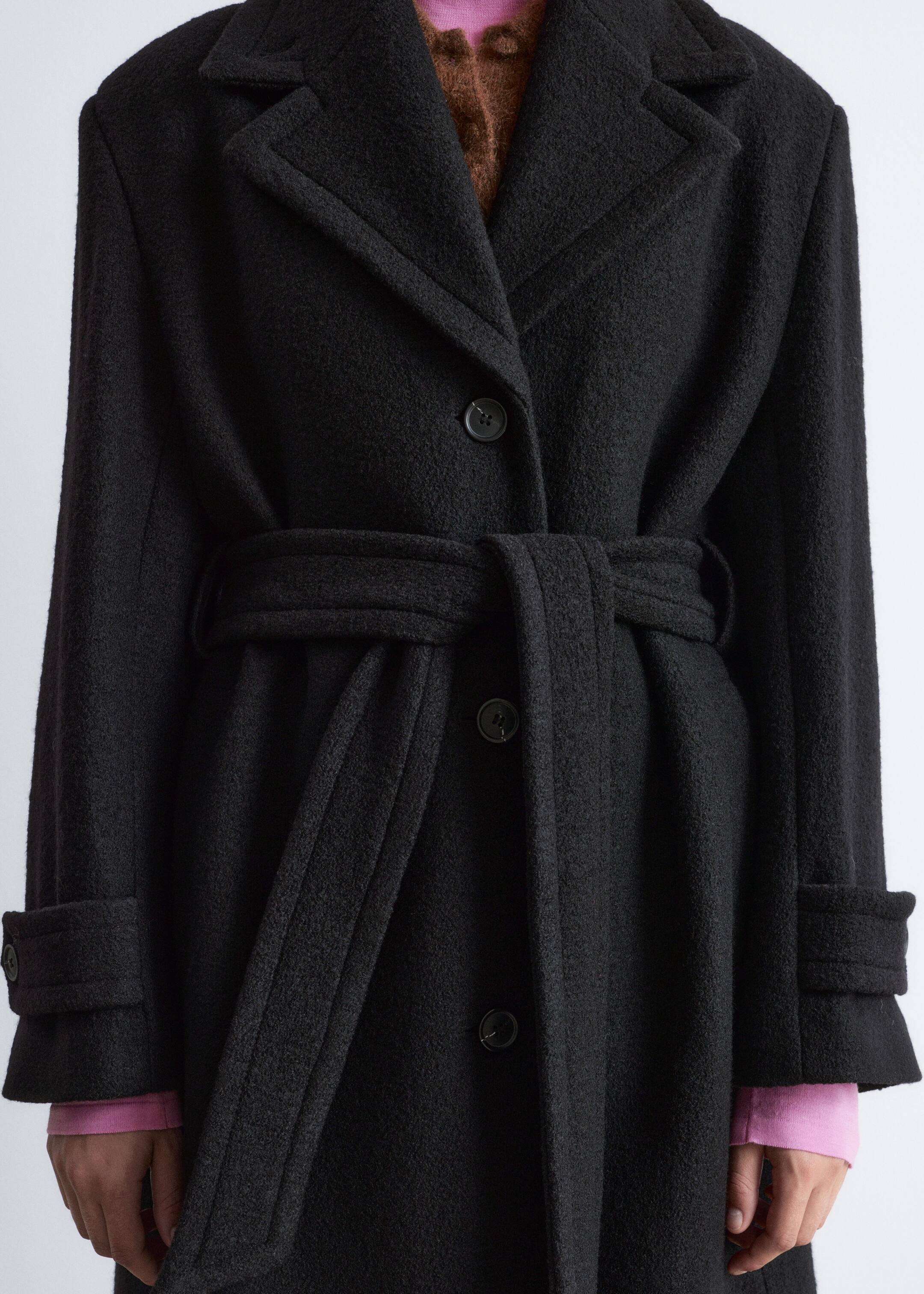 Agrandir l'image: Un manteau en laine brossée noire est présenté de face, avec des revers crantés, une fermeture à boutons simple et une ceinture en tissu qui serre la taille. Les manches longues se terminent par des poignets larges et retroussés, et un haut rose à manches longues ainsi qu'un haut texturé marron sont portés en dessous.