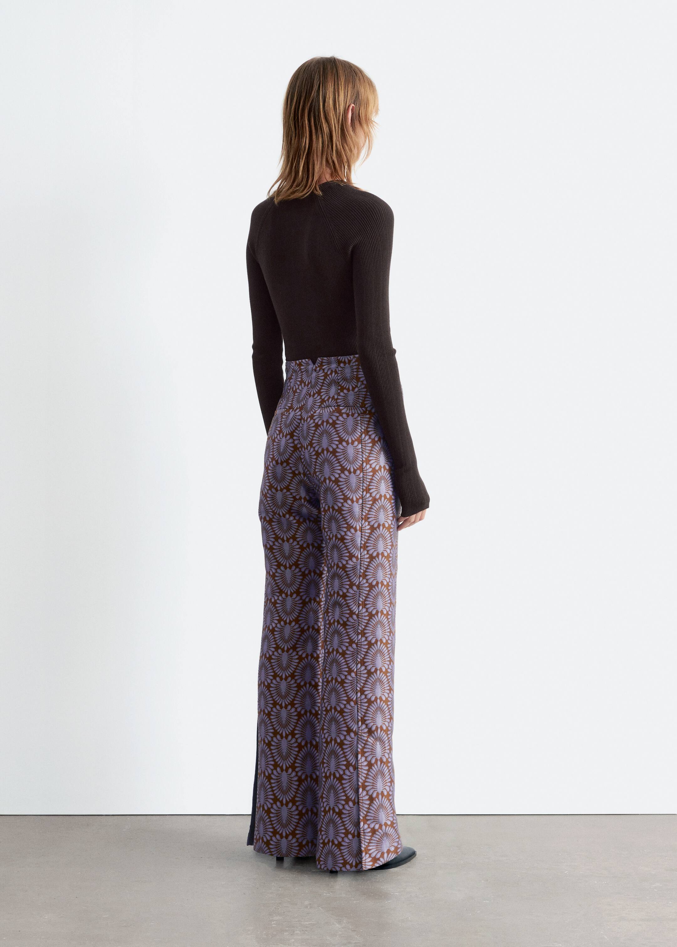 View larger image: High-waisted, wide-leg pants are featuring a repeating pattern of light purple stylized fan shapes on a brown background, seen from a back, slightly angled view. A dark brown ribbed long-sleeve top is being worn, tucked into the pants.