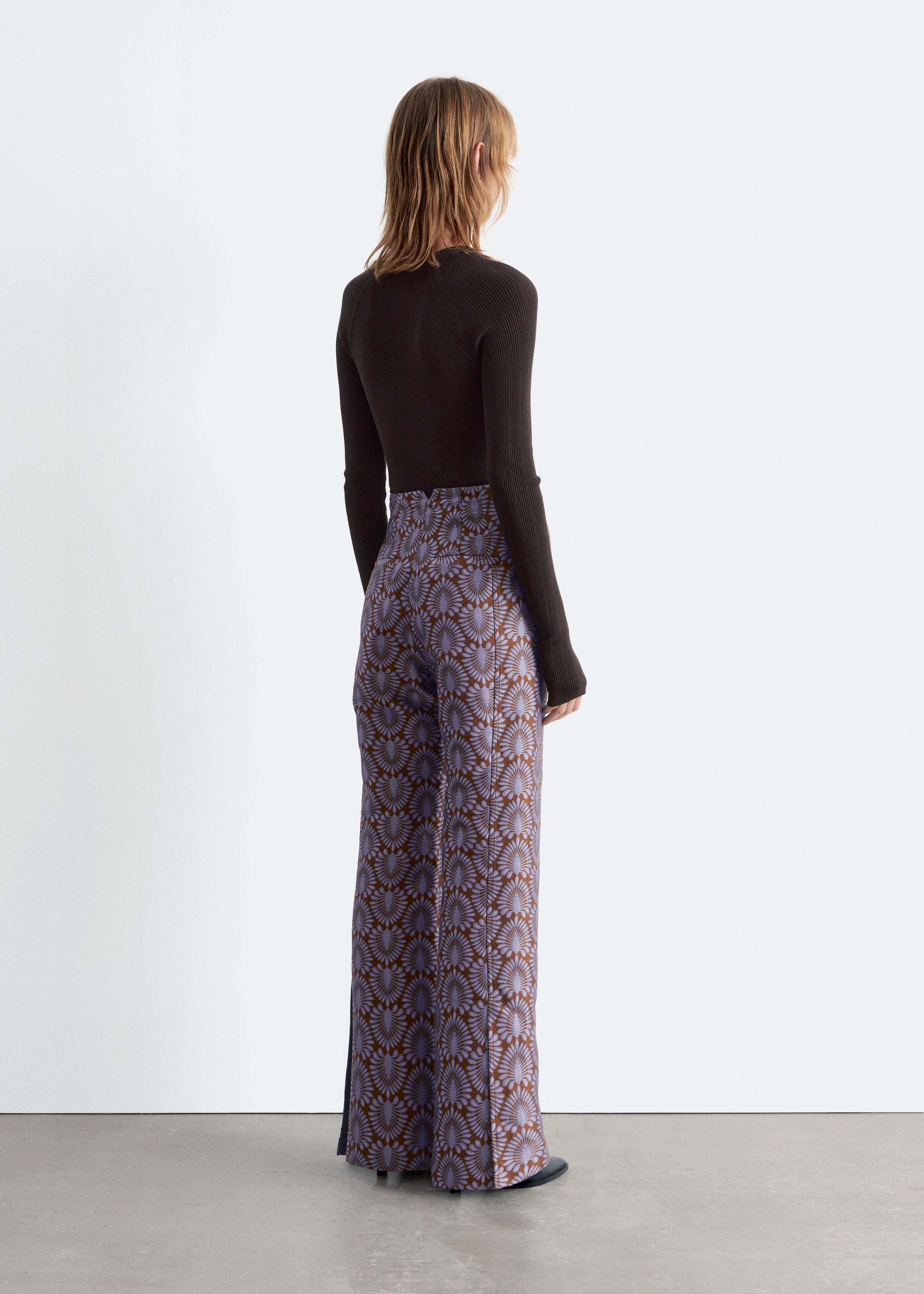Visualizza immagine più grande: Pantaloni a vita alta e gamba larga presentano un motivo ripetuto di forme di ventaglio stilizzate color viola chiaro su uno sfondo marrone, visti da dietro, con una leggera angolazione. Una maglia a maniche lunghe a coste di colore marrone scuro è indossata, infilata nei pantaloni.