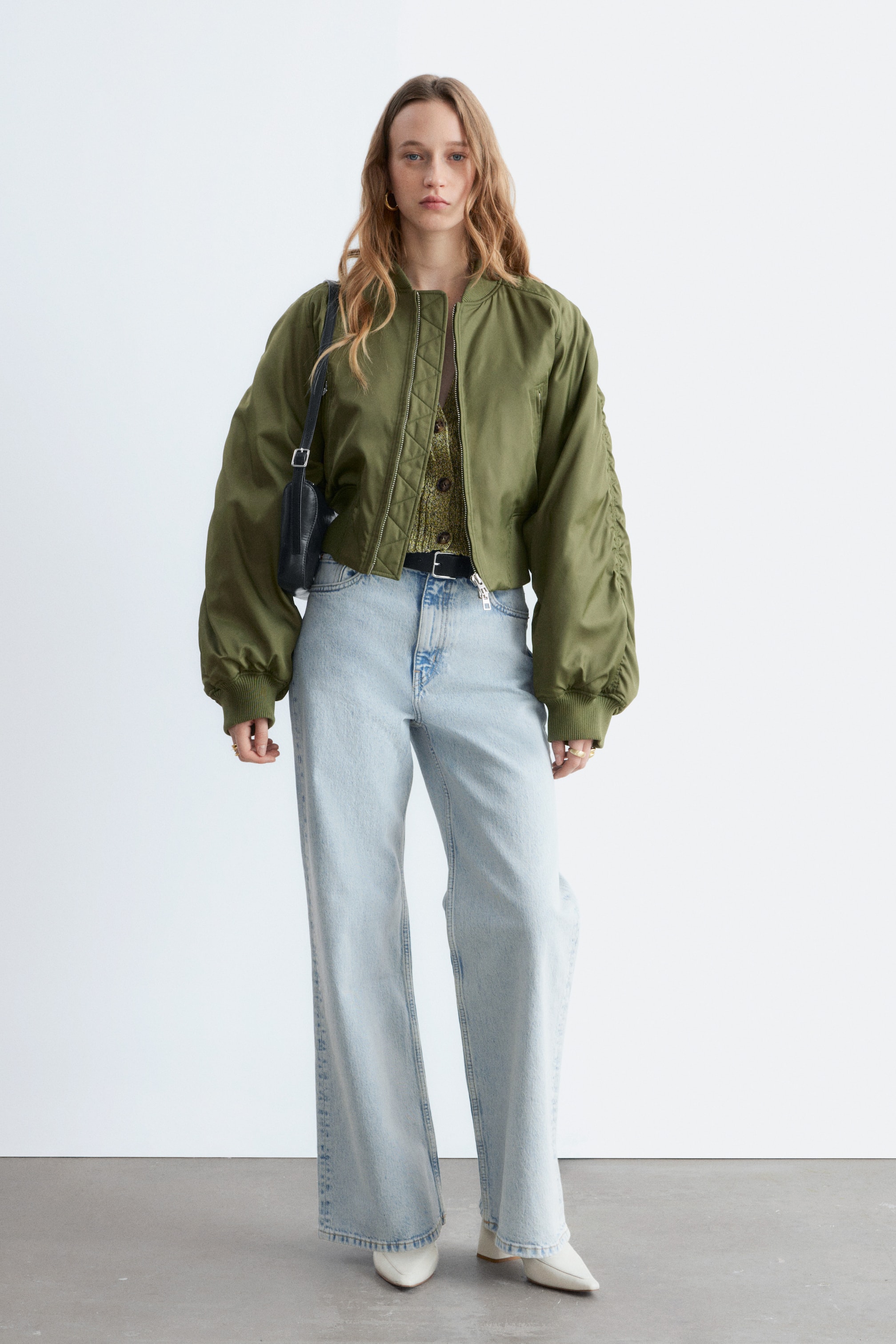 Ver imagen más grande: Se presenta una chaqueta bomber corta de color verde oliva vista desde el frente, que cuenta con un cuello alto acanalado, cierre de cremallera frontal y un dobladillo acanalado. Muestra mangas largas y voluminosas con detalles fruncidos en la parte superior del brazo y puños acanalados, y se lleva abierta sobre un cárdigan de punto verde.