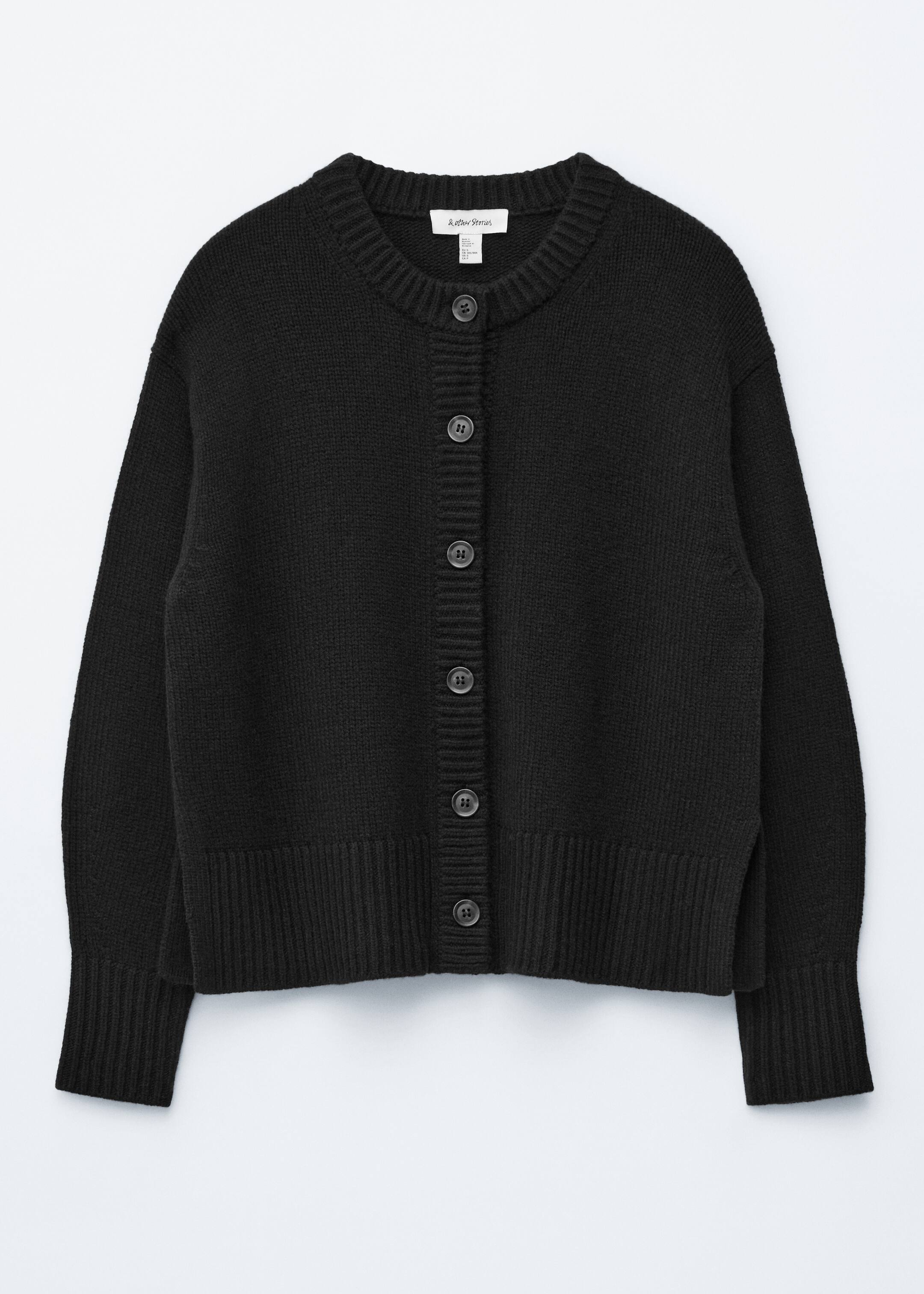 Agrandir l'image: Un cardigan en tricot noir est présenté de face, avec un col rond côtelé et six boutons circulaires sombres au centre. Il présente des manches longues avec des poignets côtelés et un ourlet côtelé avec de petites fentes latérales, indiquant une coupe décontractée.