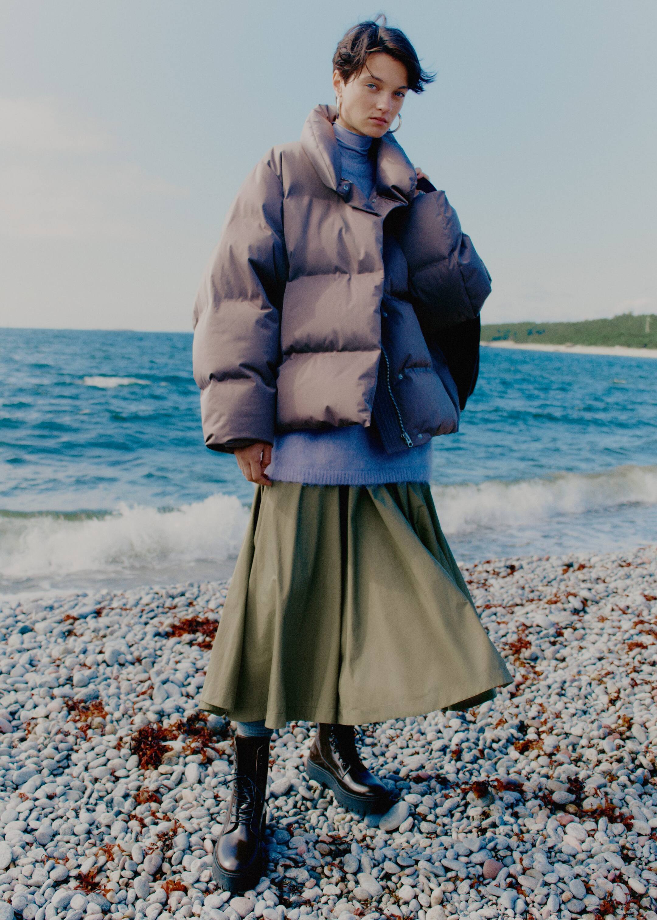 Größeres Bild anzeigen: Eine dunkelgraue Daunenjacke wird von einer Person getragen, die leicht nach rechts schaut. Sie zeigt eine voluminöse, übergroße Silhouette mit horizontalen Steppnähten, einem hochstehenden Kragen und einem sichtbaren, teilweise offenen Frontreißverschluss. Die Jacke wird offen getragen und offenbart einen hellen lila Rollkragenpullover darunter.