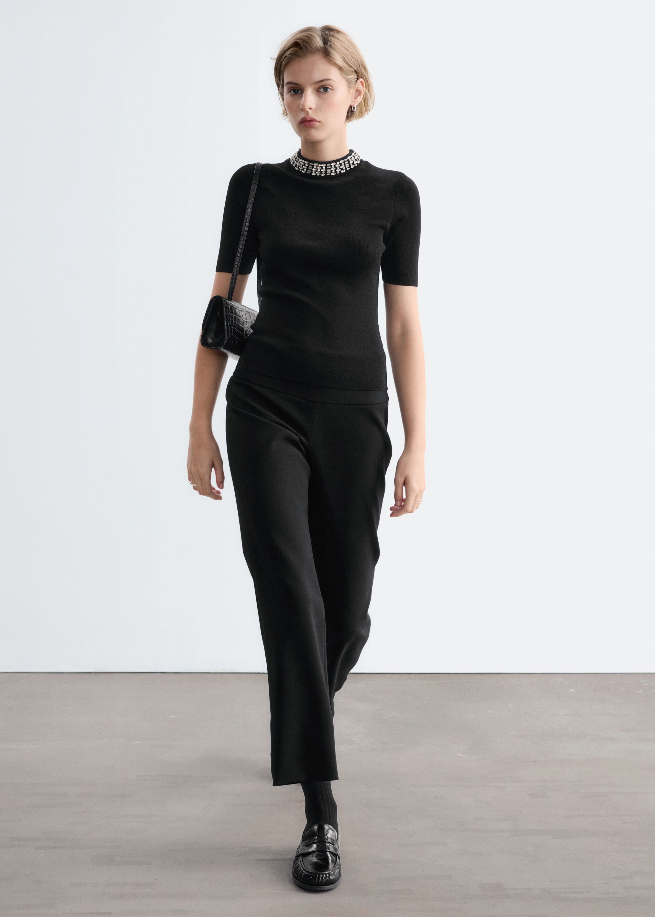 A black, fitted, short-sleeved top featuring a mock neck collar embellished with rows of silver rhinestone beads is worn by the model. The top is styled with black trousers and a black shoulder bag.