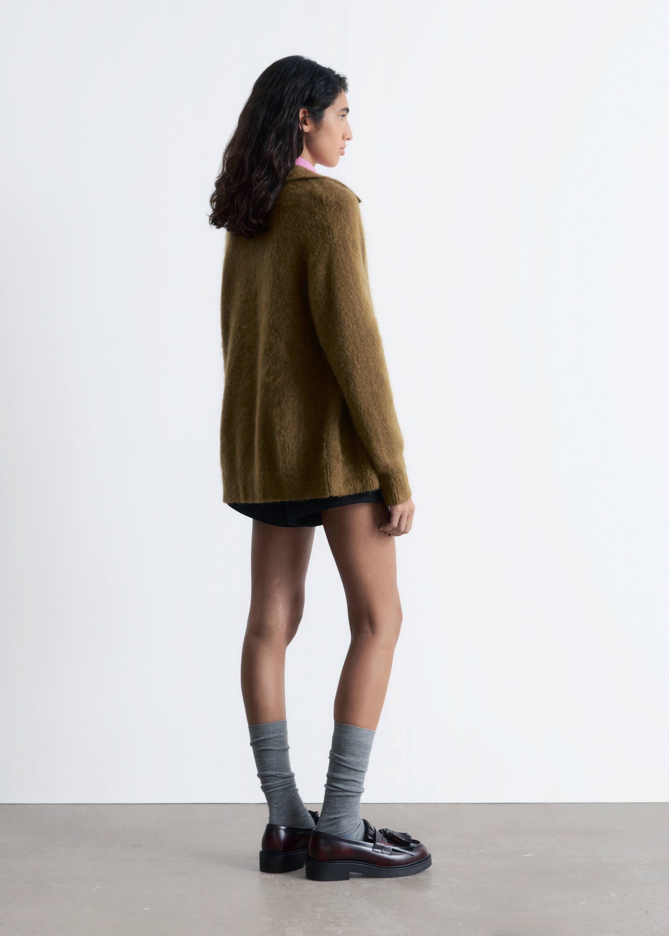 Grösseres Bild ansehen: Eine gedämpfte olivgrüne, flauschige Strickjacke wird getragen, von der Seite betrachtet, die ihre lockere Passform und verlängerte Länge zeigt. Ein leuchtend pinkes Oberteil lugt unter dem Kragen hervor, und schwarze Shorts sind unter dem Saum der Strickjacke sichtbar.