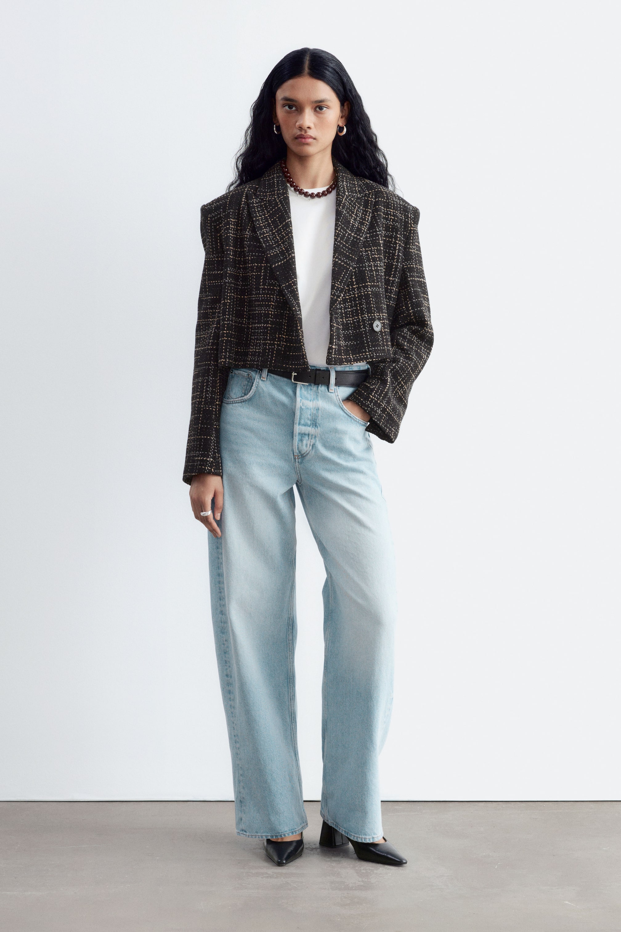 Agrandir l'image: Un jean bleu délavé clair, doté d’une taille mi-haute et d’une silhouette oversize à jambe barrel, est porté par le mannequin. Le jean est associé à une ceinture noire, un t-shirt blanc et un blazer court à motif foncé, tandis que le mannequin repose une main dans une poche avant.
