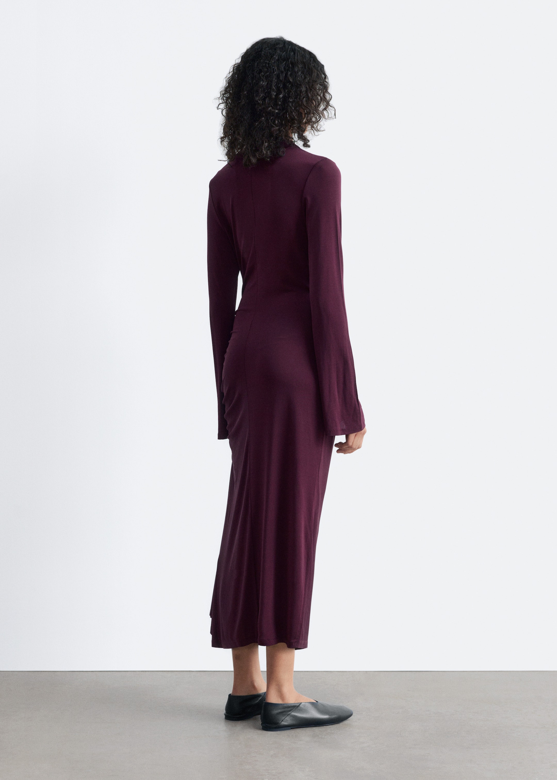 Größeres Bild anzeigen: Ein bordeauxrotes Midikleid wird von dem Model getragen, das mit dem Rücken zum Betrachter steht. Das figurbetonte Kleid ist mit einem Stehkragen, langen ausgestellten Ärmeln und einer gerafften Seitenaht, die in einen drapierten Saum übergeht, gestylt. Das Model trägt auch schwarze flache Schuhe.