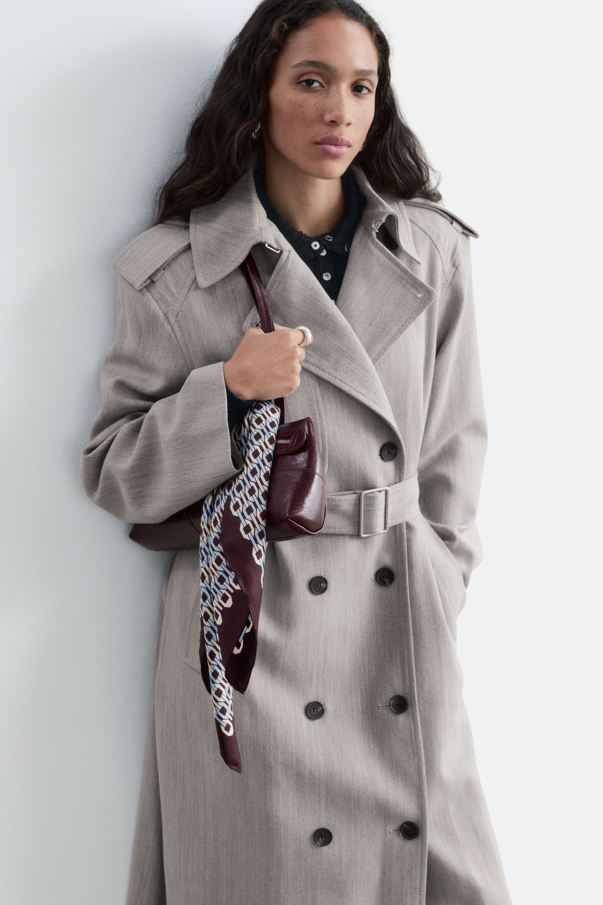 Visualizza immagine più grande: La modella indossa un trench doppiopetto in misto cotone e lana di colore grigio chiaro, impreziosito da una cintura in vita con fibbia. Alla tracolla di una borsa a spalla in pelle rosso scuro, portata dalla modella, è annodato un leggero foulard in seta con stampa a catena all-over.