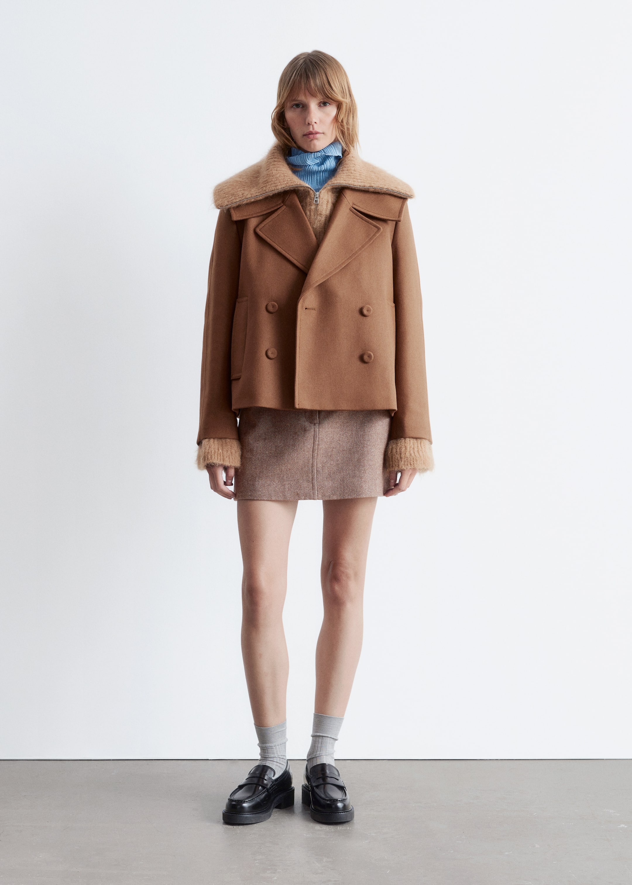 Agrandir l'image: Une veste camel à double boutonnage avec de larges revers en pointe, un col contrastant en fourrure marron clair et des poignets assortis est portée par le mannequin. La veste a une fermeture à boutons et deux poches passepoilées à l'avant, et est superposée sur un col roulé bleu clair et un pull marron clair.