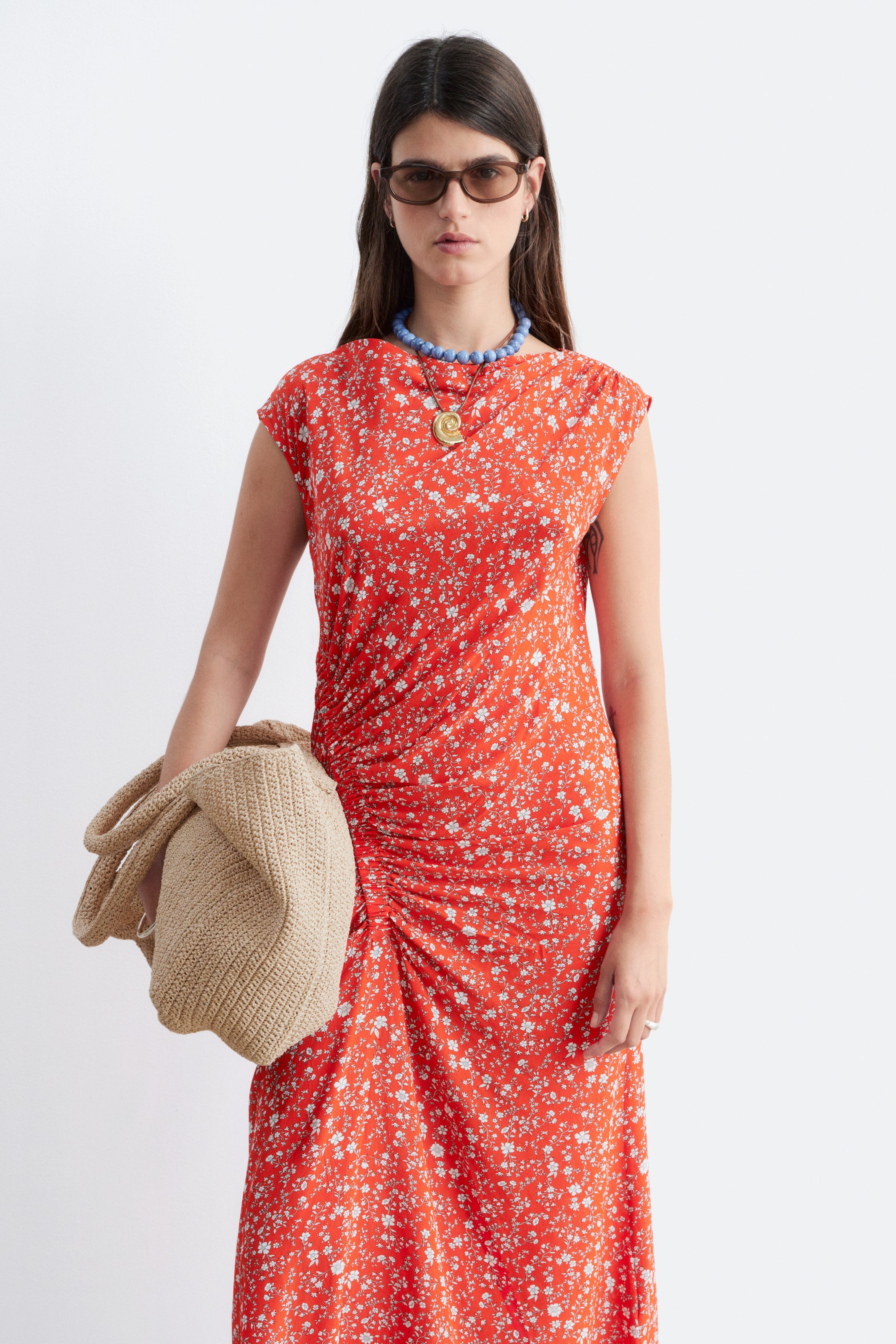 Grotere afbeelding bekijken: De vooraanzicht van de rode jurk toont een wit bloemenpatroon, een boothals en kapmouwtjes. Plooien zijn zichtbaar langs de linkerkant van de jurk, wat een gedrapeerd effect creëert.