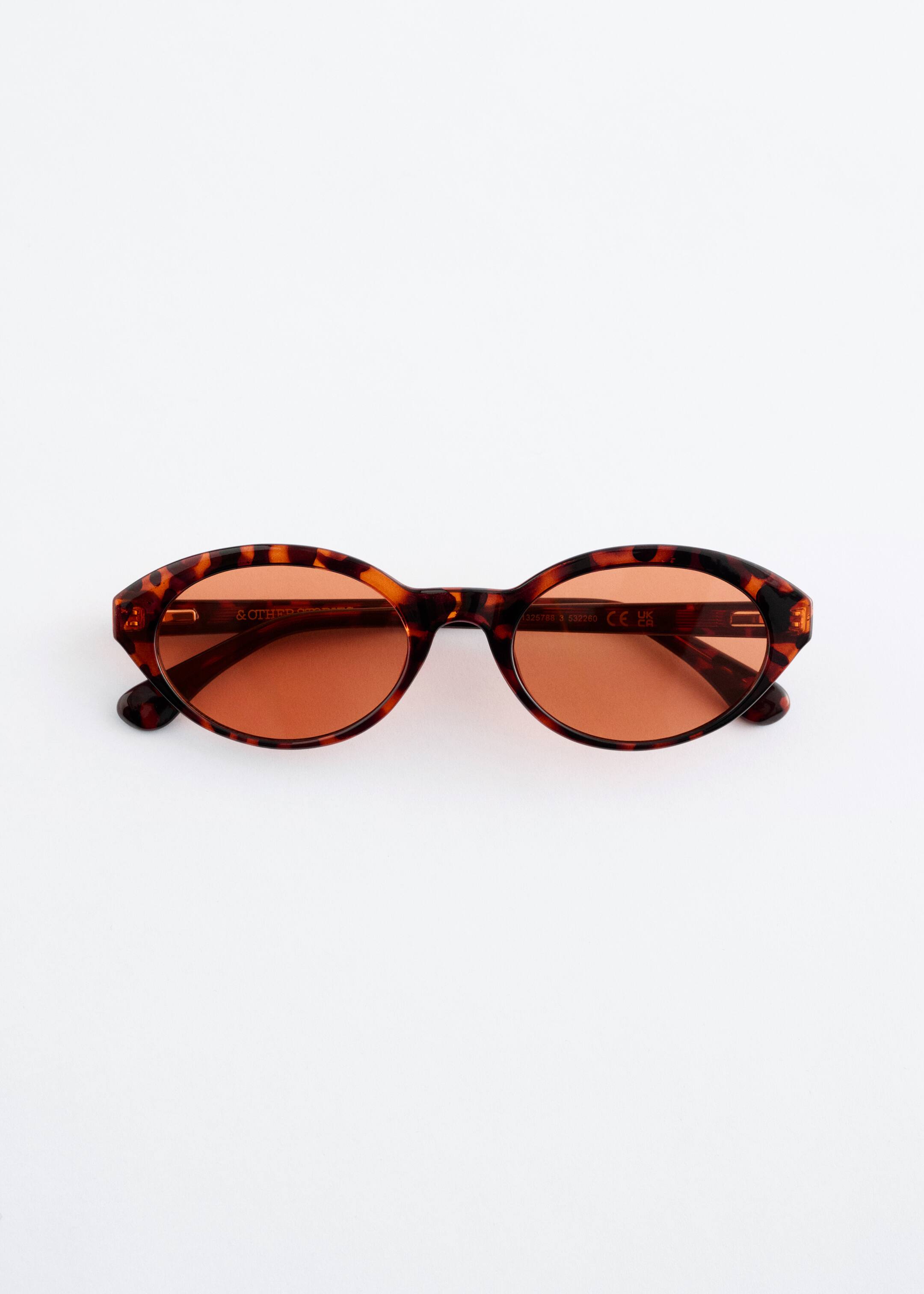 Zobrazit větší obrázek: Pár slunečních brýlí představuje FRONT, s úzkými, oválnými obroučkami s hnědo-černým želvovinovým vzorem. Obroučky obklopují oválné, oranžově tónované čočky.