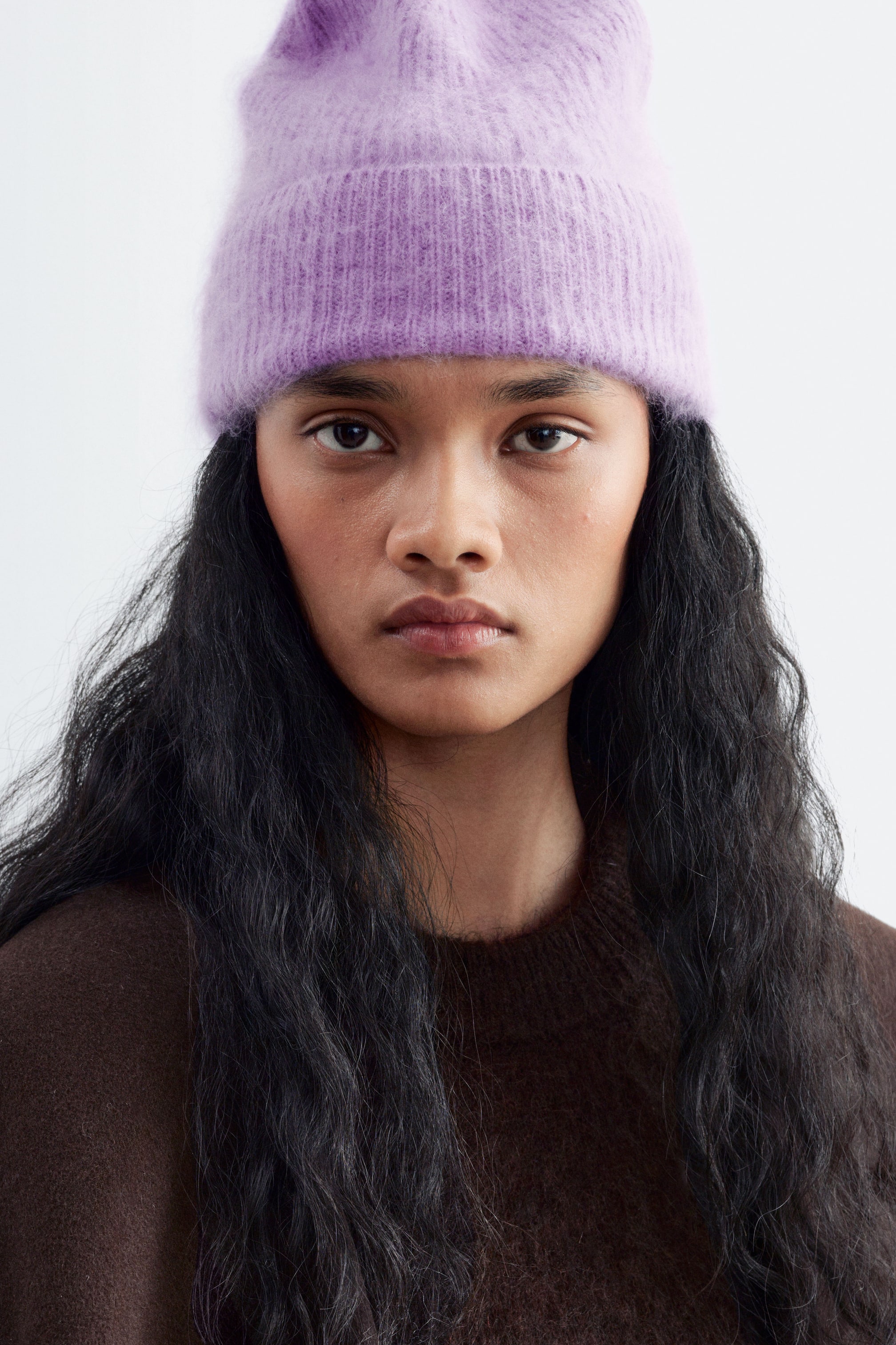 Größeres Bild ansehen: Eine hellviolette, flauschige Strickmütze mit umgeschlagenem Bund wird von dem Model getragen. Die Mütze wird mit einem dunkelbraunen Rundhalspullover kombiniert.