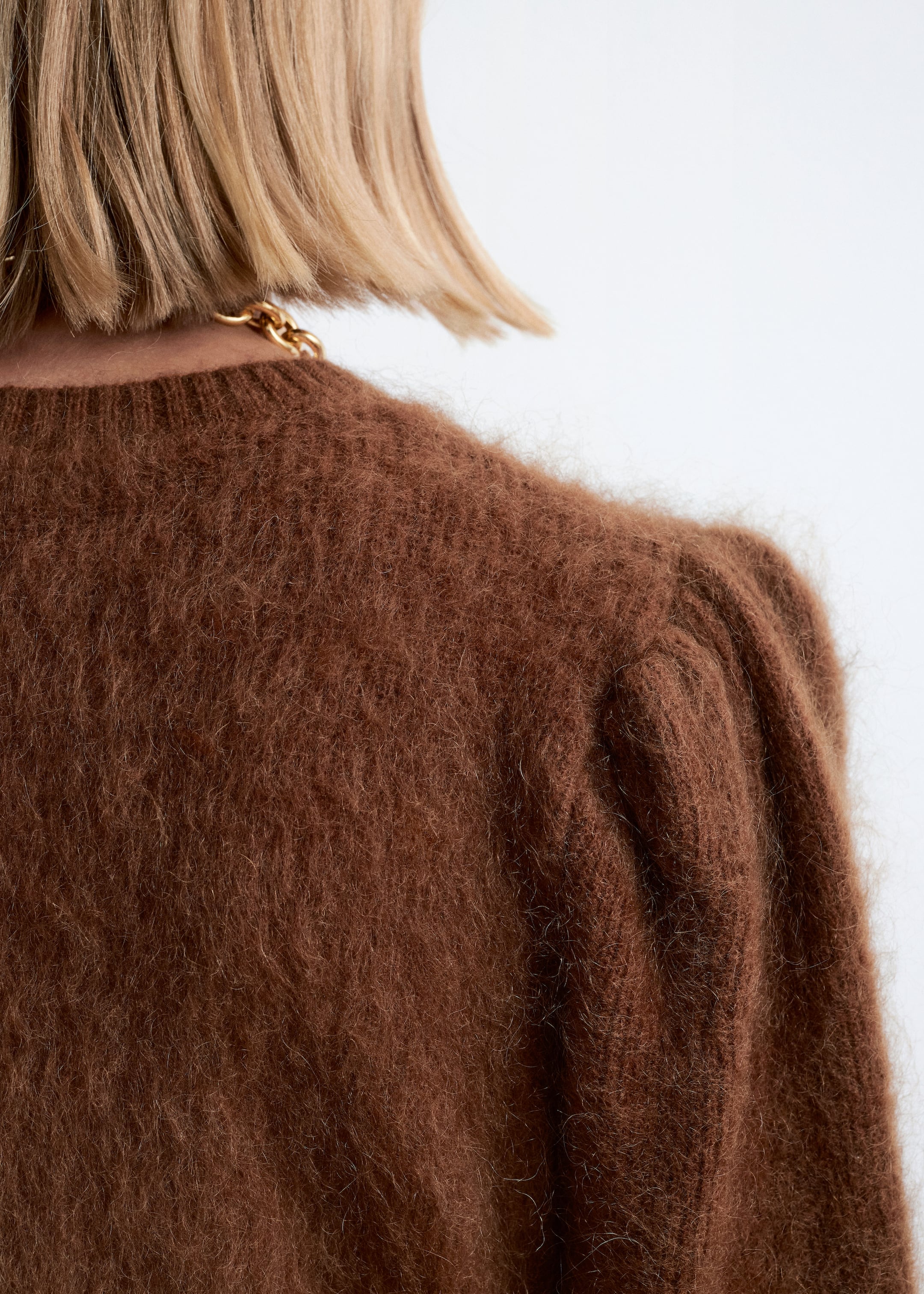 Visualizza immagine più grande: Un maglione marrone ruggine viene visto da dietro, mettendo in risalto la sua texture pelosa e lavorata a maglia spazzolata e un sottile dettaglio di manica a sbuffo sulla spalla destra. Una catena d'oro è appoggiata sullo scollo a girocollo del maglione.