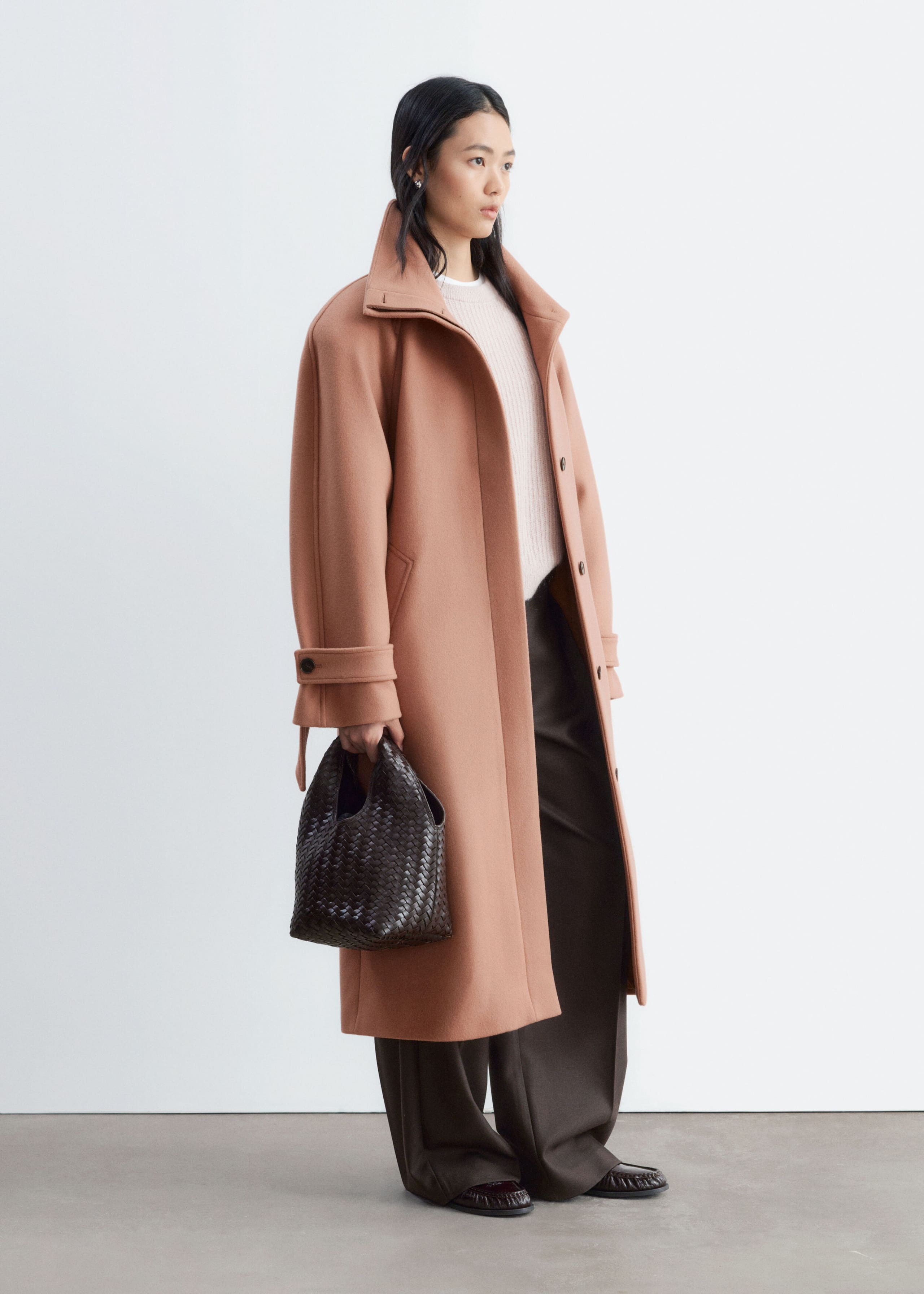 A long, peach-coloured coat with a high collar and structured shoulders is worn open by the model over a light-coloured knit sweater and dark, wide-leg trousers. The model is also carrying a dark brown woven handbag at their side.