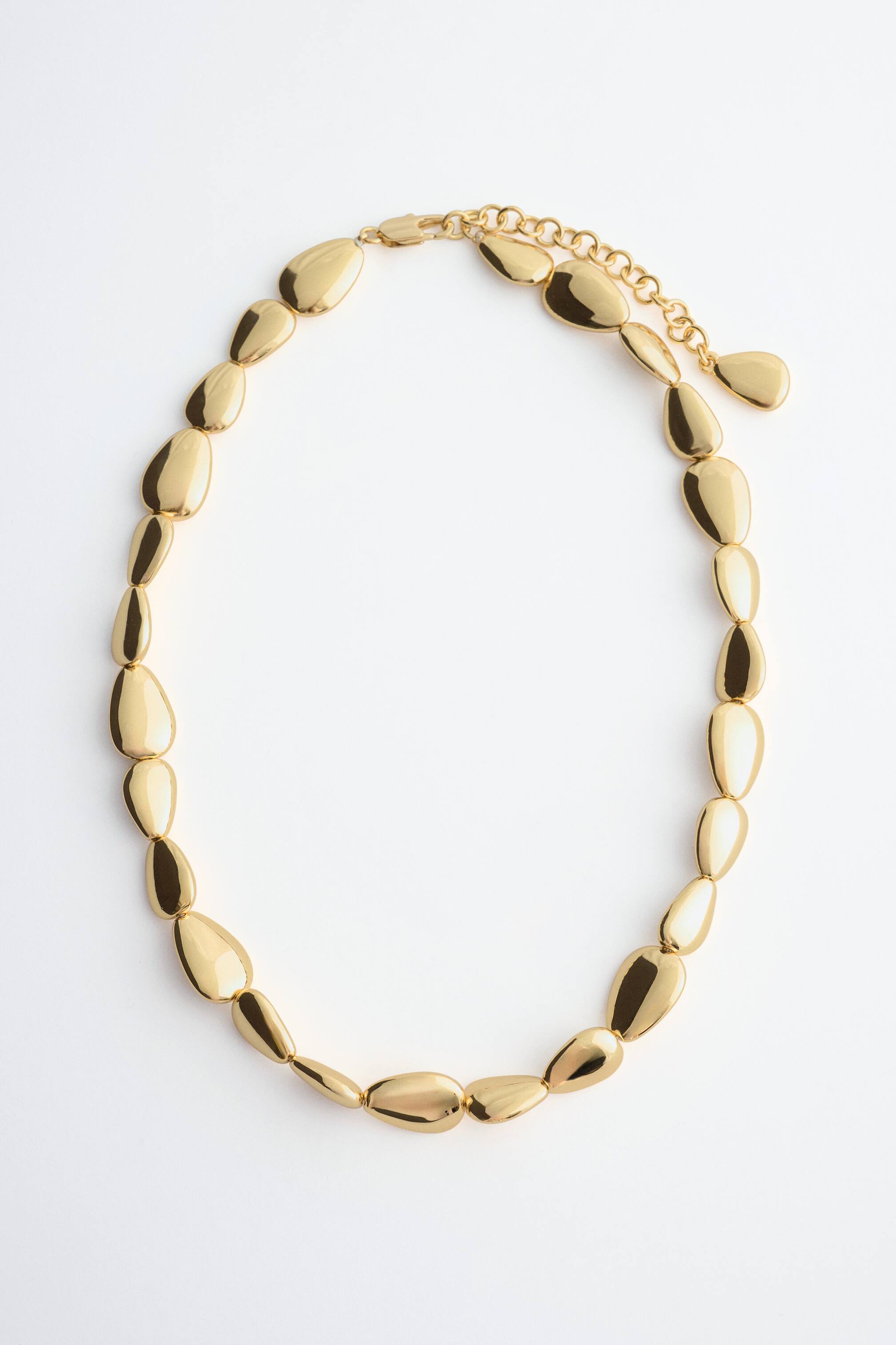 Agrandir l'image: Un collier doré poli est disposé à plat, composé d’une série de maillons lisses, aux formes organiques, rappelant des galets polis. Le collier est doté d’une chaîne d’extension réglable et d’un fermoir mousqueton.