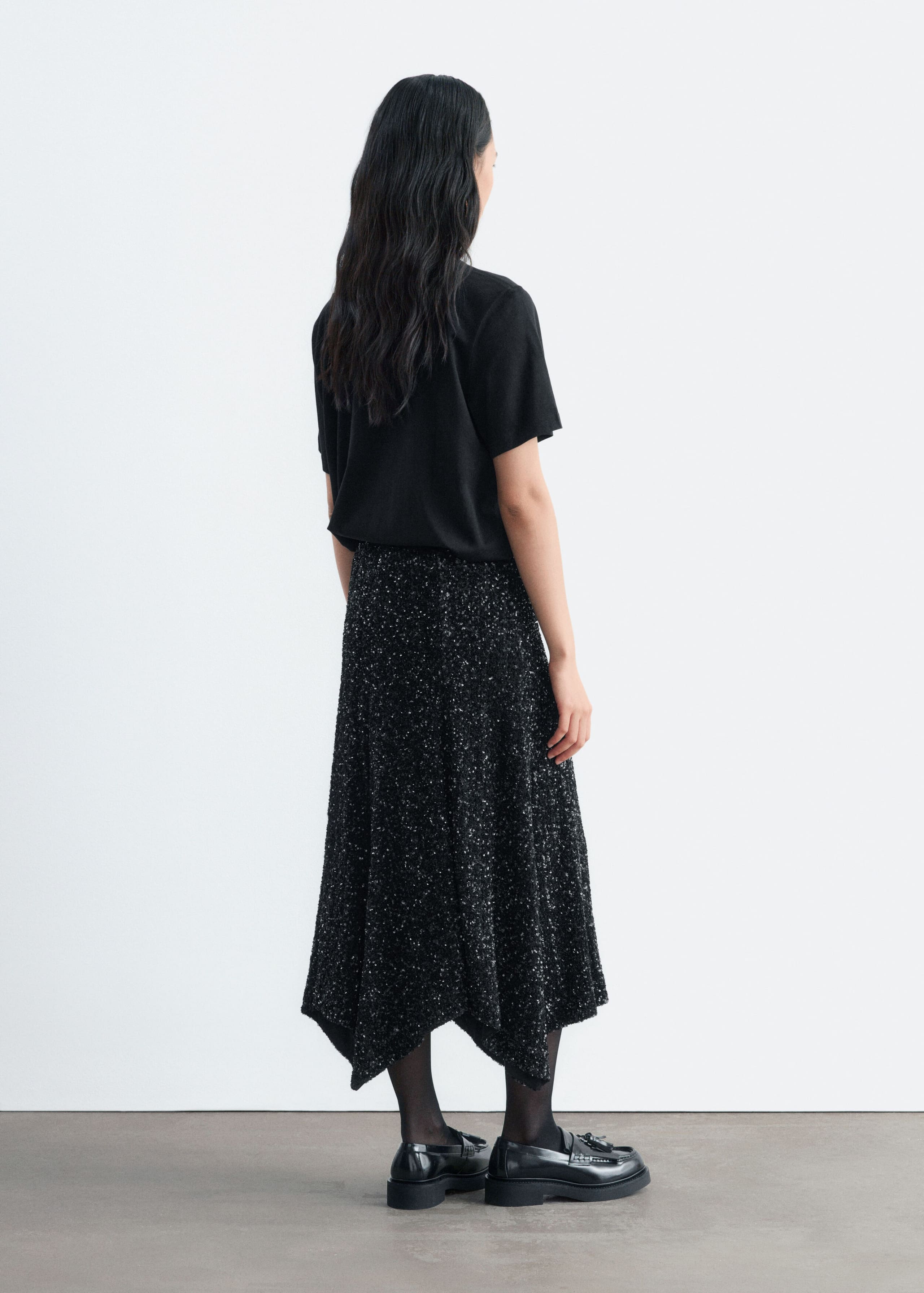 The model is wearing a black sequined mesh skirt with an asymmetric hem and is shown in a three-quarter back view. The skirt is styled with a tucked-in black short-sleeve T-shirt, sheer black tights, and black patent loafers.