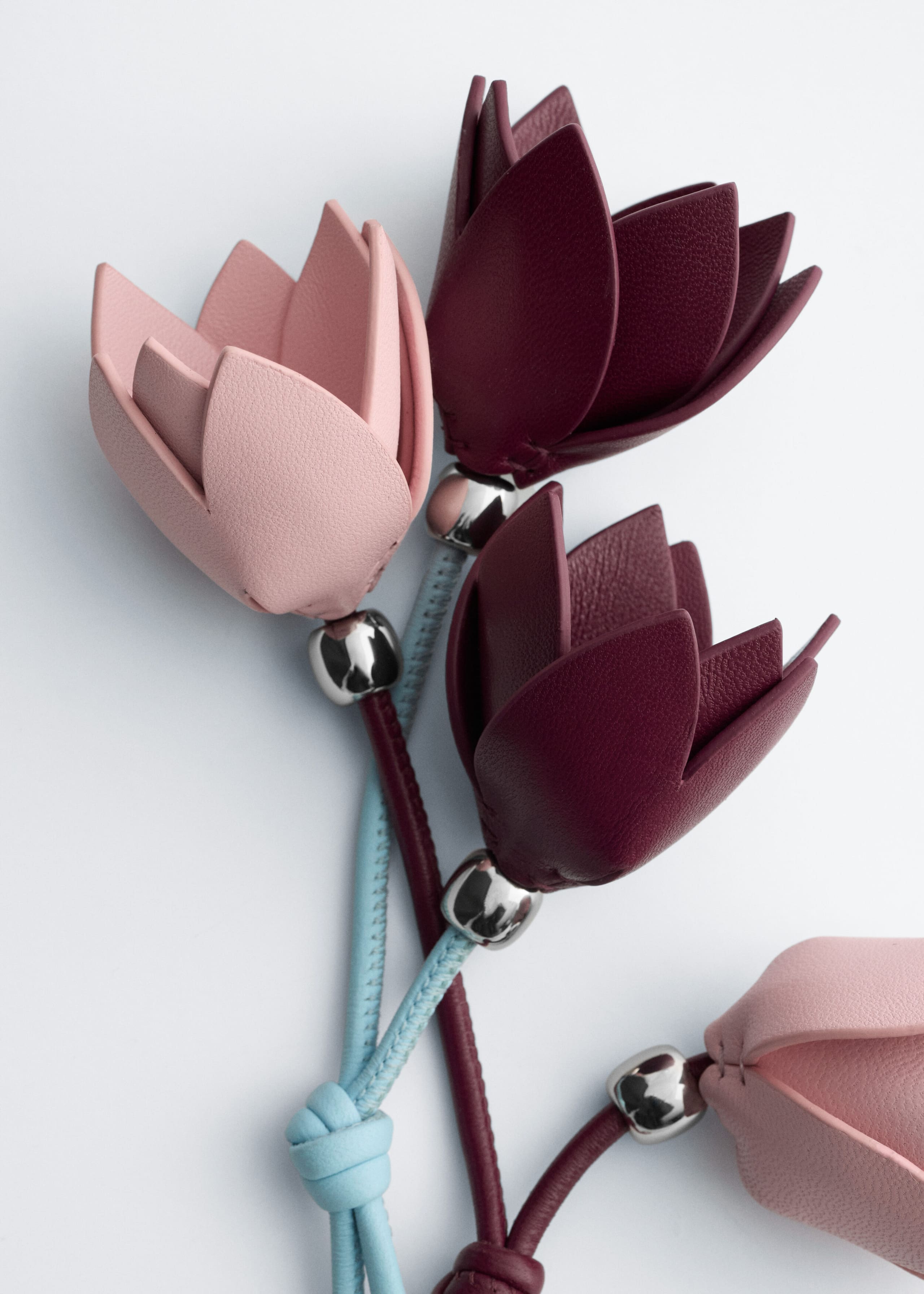 Die Flower Bag Charms zeigen tulpenförmige Lederblütenköpfe in Hellrosa und tiefem Burgunderrot, gefertigt mit übereinandergelegten Blütenblättern und akzentuiert durch silberfarbene Metallperlen. Dünne Lederkordeln in Hellblau und Burgunderrot verlaufen von den Blütenbasen nach unten und sind in sauberen Knoten zusammengebunden.