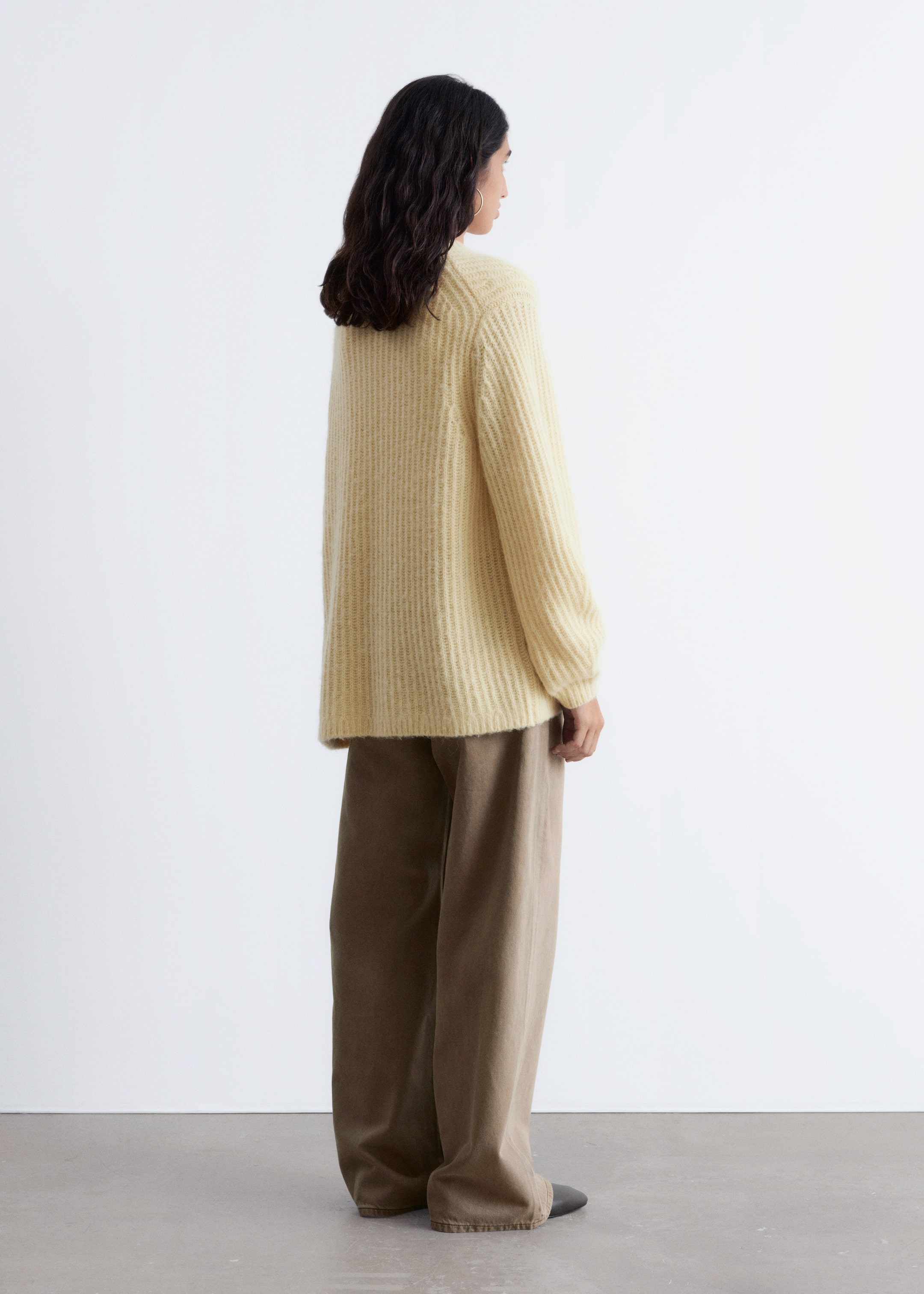 Visualizza immagine più grande: Un maglione a maglia a coste di colore giallo chiaro viene presentato in un profilo laterale, mettendo in risalto la sua vestibilità rilassata e oversize e le maniche lunghe, leggermente voluminose che si restringono ai polsi. Il maglione è abbinato a pantaloni marroni a gamba larga.