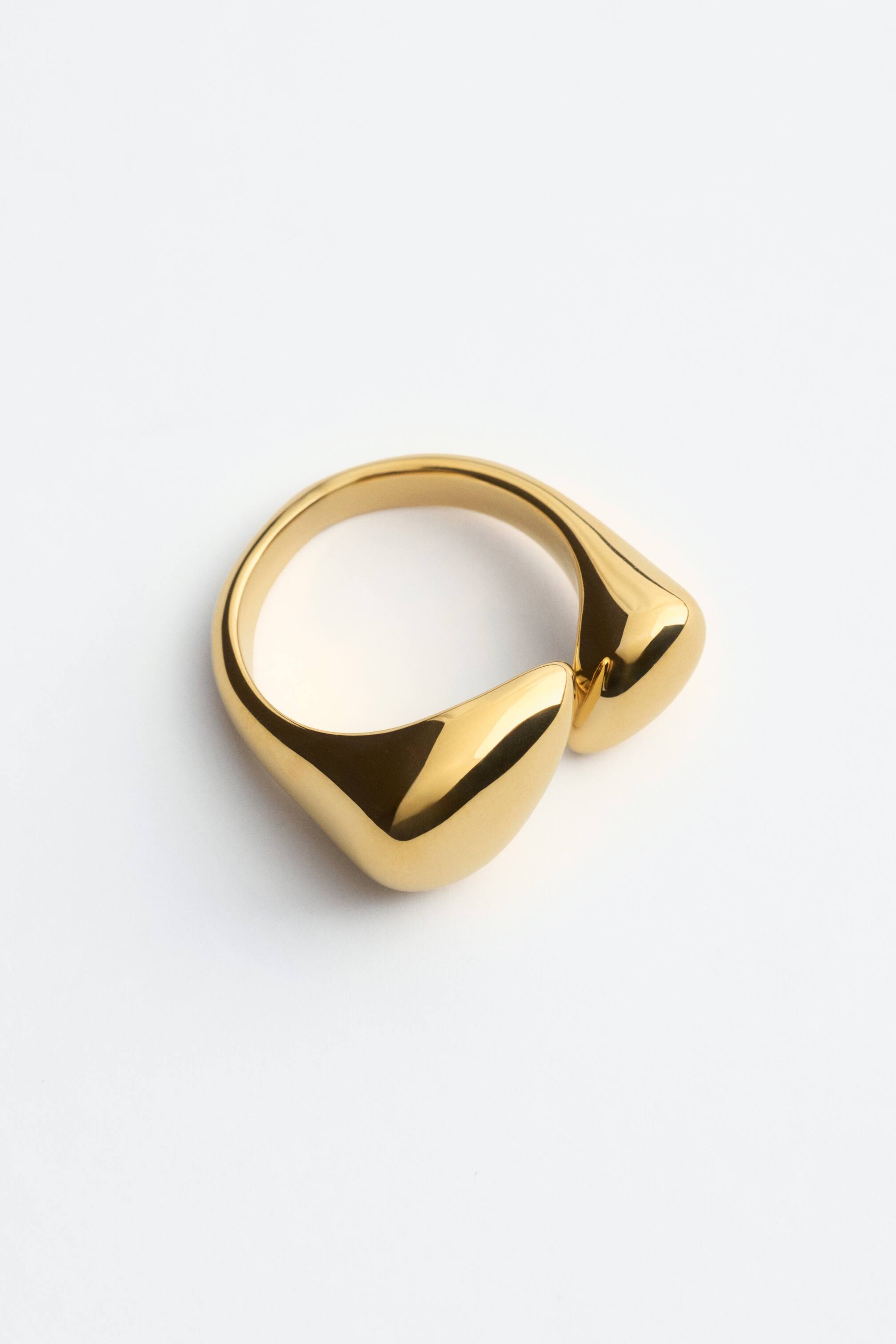 Grösseres Bild ansehen: Ein polierter Messingring zeigt aus einer schrägen Vorderansicht eine glatte, reflektierende Oberfläche und eine fließende, skulpturale Silhouette. Das offene Design an der Vorderseite zeigt zwei voluminöse, abgerundete Formen, die sich einander entgegenbiegen.
