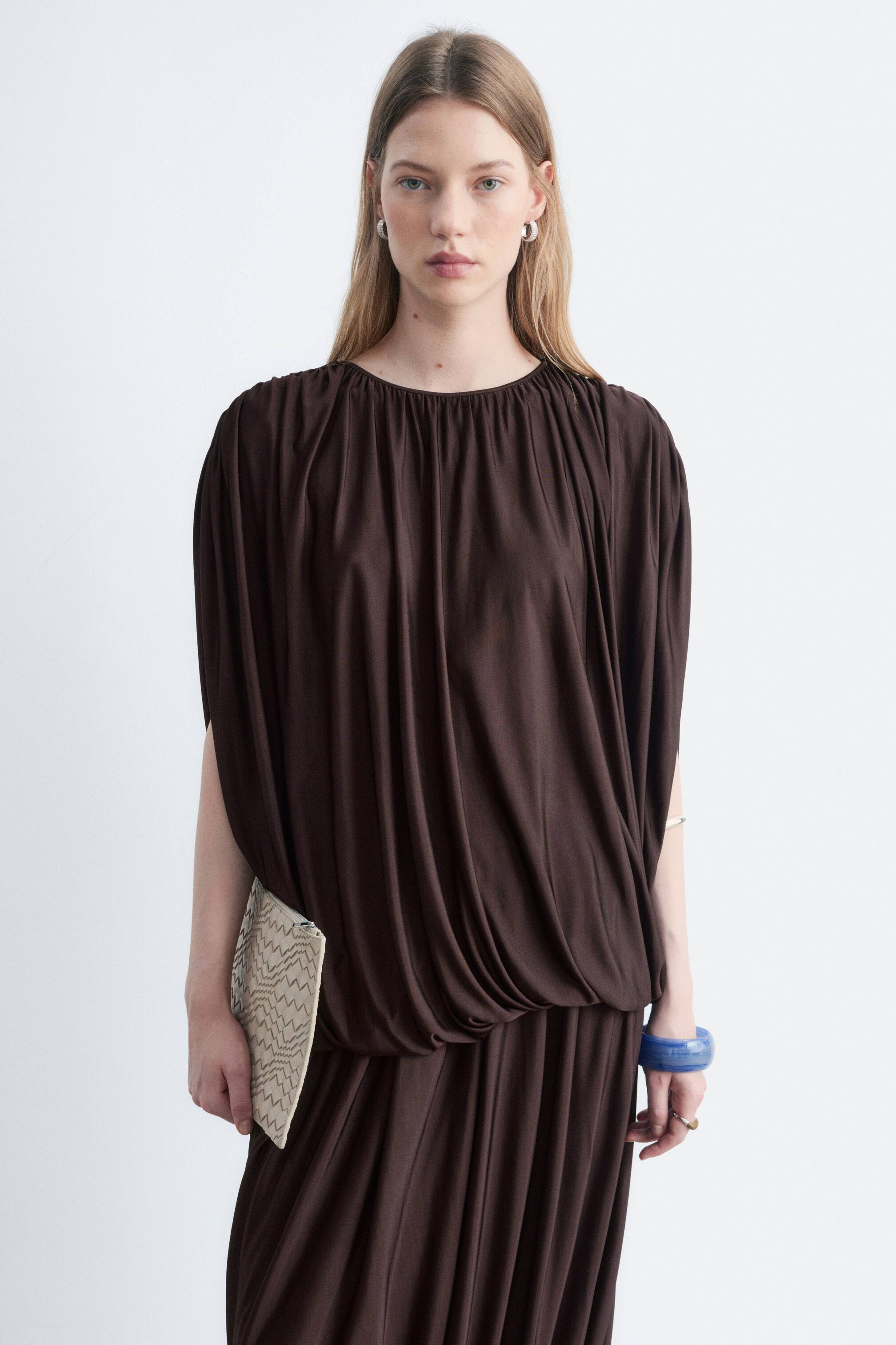 Visualizza l'immagine ingrandita: La modella indossa un top in jersey marrone scuro senza maniche, con scollo tondo arricciato e ampie drappeggiature sul corpetto. La modella tiene una clutch con motivo a contrasto sul top, abbinato a una gonna plissettata coordinata.