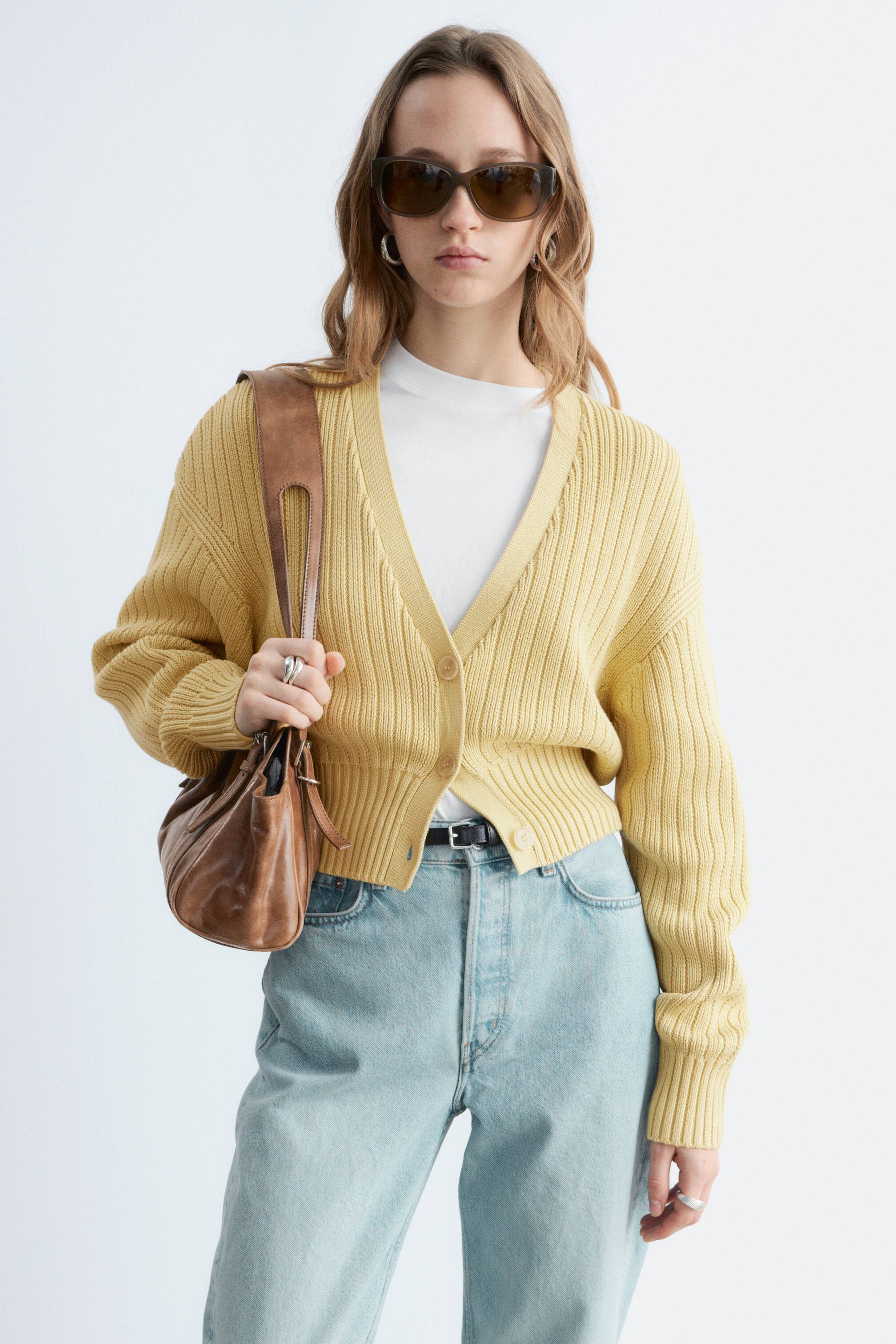 Ver imagem maior: Um cardigan amarelo de malha canelada é apresentado de frente, com um decote em V, três botões na frente, mangas compridas com punhos canelados e uma bainha canelada e curta. Está sobreposto a uma t-shirt branca.