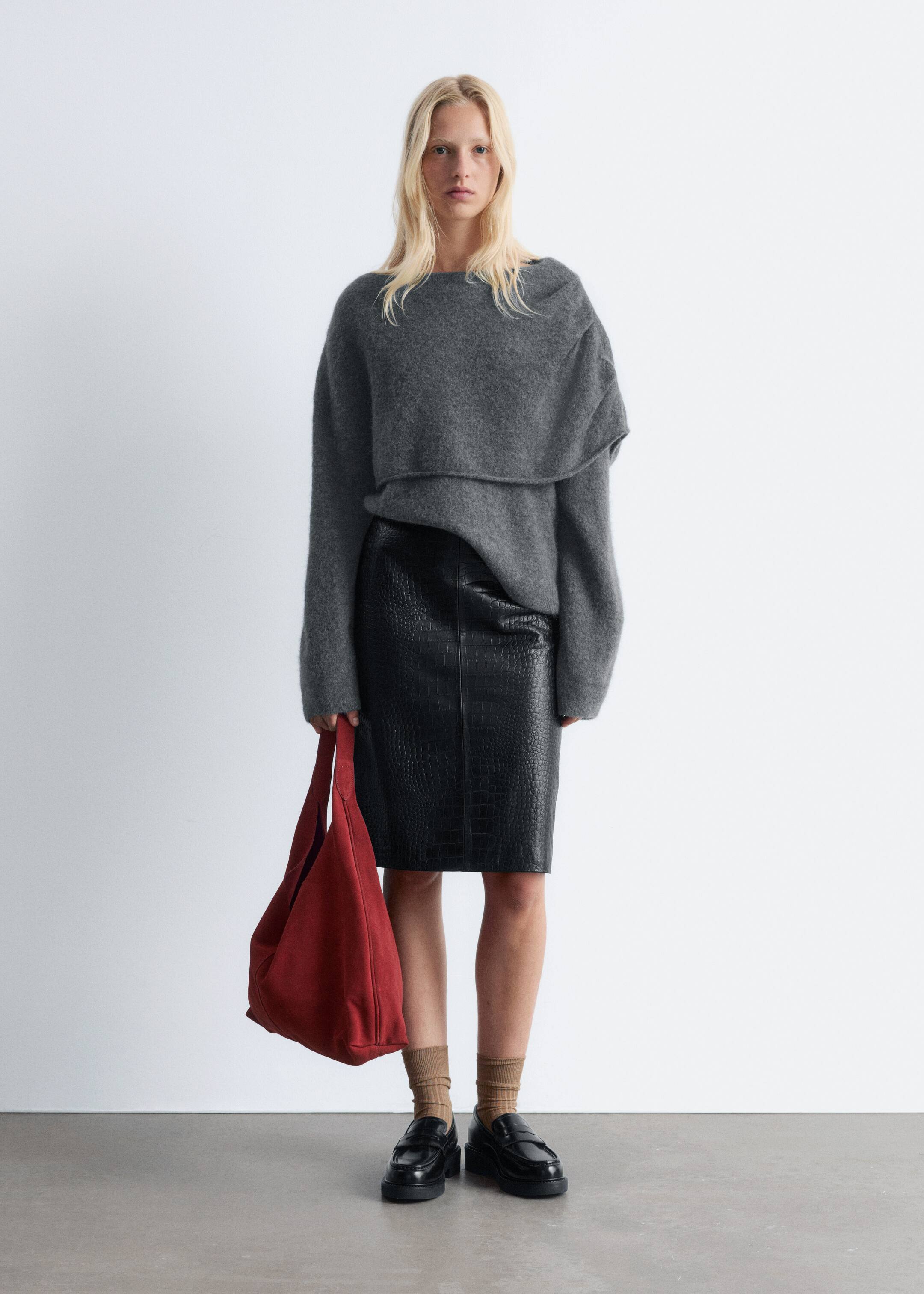 Ver imagem maior: Uma modelo está voltada para a frente, vestindo uma camisola cinzenta oversized com decote barco e um detalhe drapeado e em camadas no peito, combinada com uma saia lápis preta com relevo de crocodilo. Uma mala hobo vermelha e desleixada está a ser segurada, e mocassins pretos robustos estão a ser usados com meias caneladas.