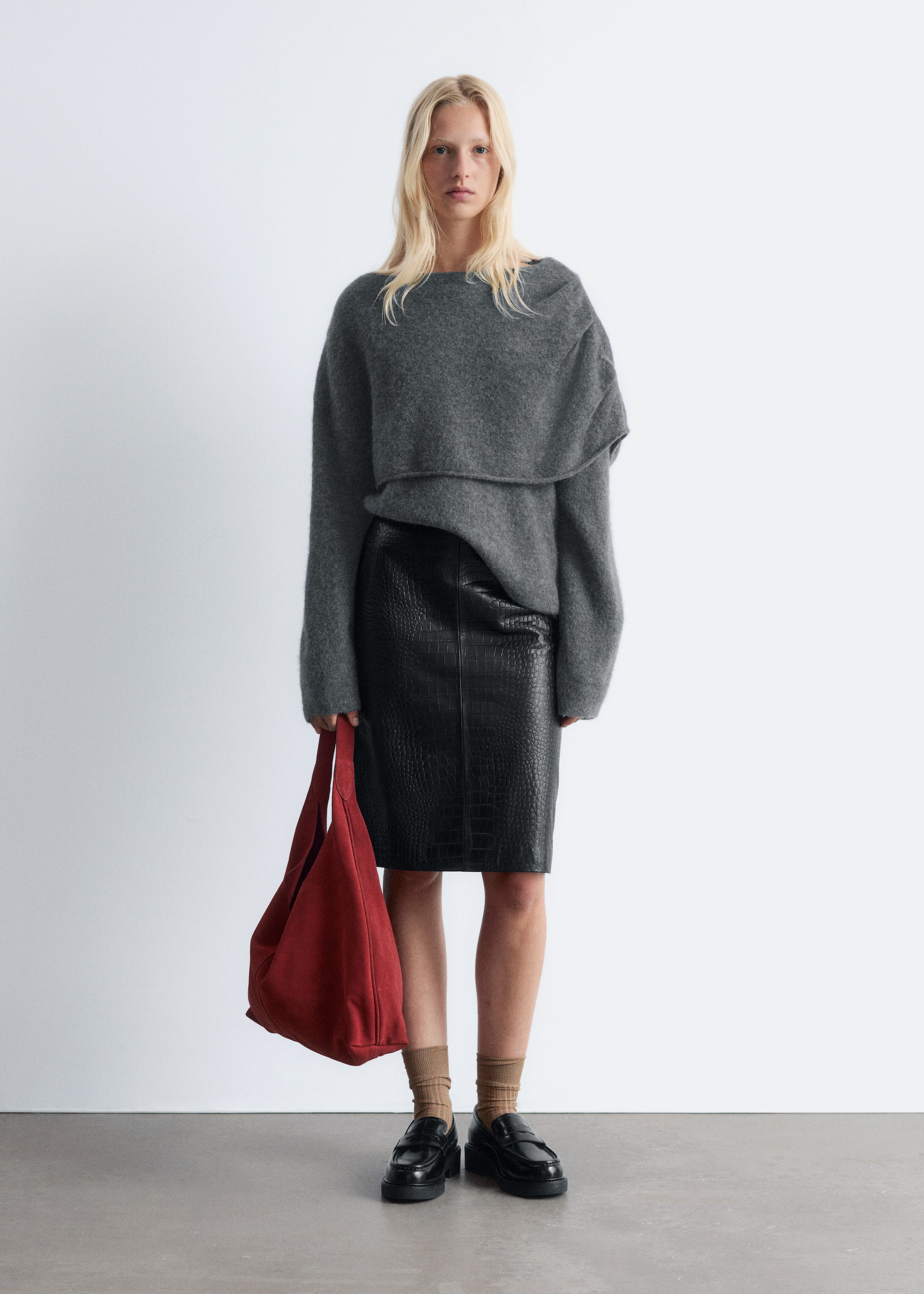 Zobacz większy obraz: Modelka stoi przodem, ma na sobie szary, oversize'owy sweter z dekoltem w łódkę i drapowanym, warstwowym detalem na klatce piersiowej, w połączeniu z czarną spódnicą ołówkową z tłoczeniem w krokodyla. Trzyma czerwoną, luźną torbę typu hobo, a na nogach ma czarne, masywne loafersy z prążkowanymi skarpetami.