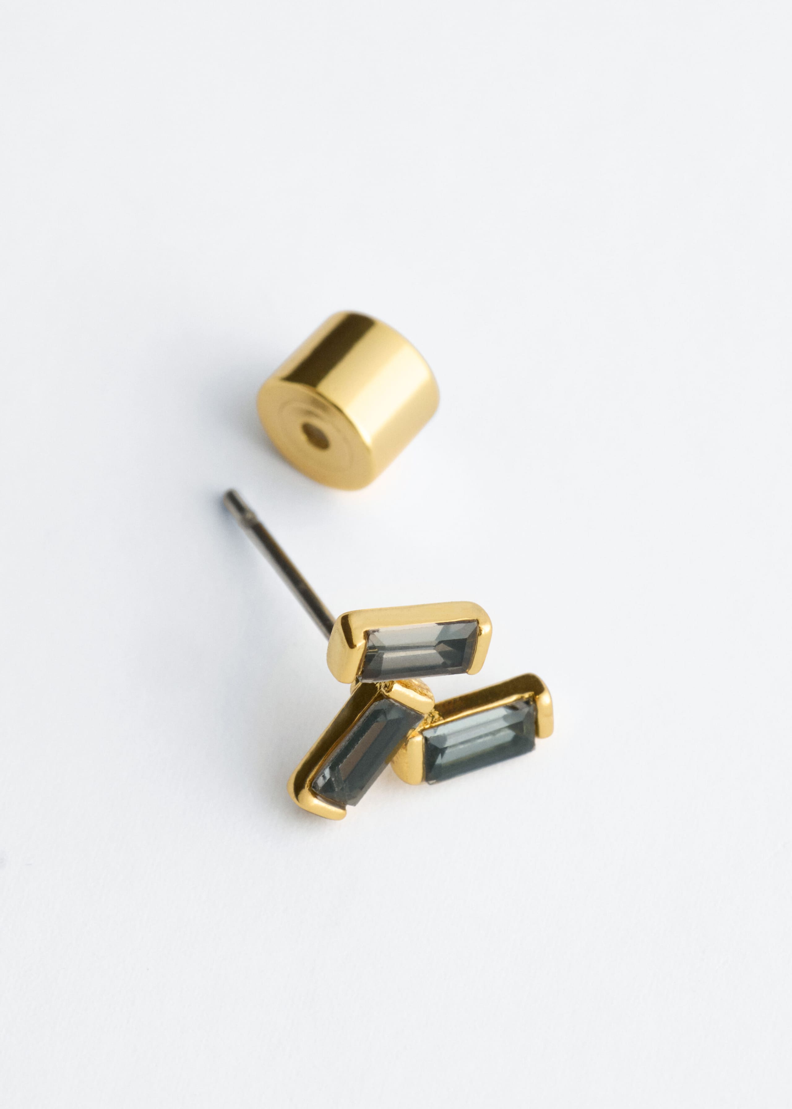 Dieser goldfarben schimmernde Ohrstecker aus Messing ist mit einer Gruppe aus drei grauen, baguettgeschliffenen Zirkonia-Steinen in Zargenfassungen versehen. Ein passender, zylindrischer Ohrstecker-Verschluss in Goldoptik liegt separat daneben und zeigt den Stift des Ohrsteckers.
