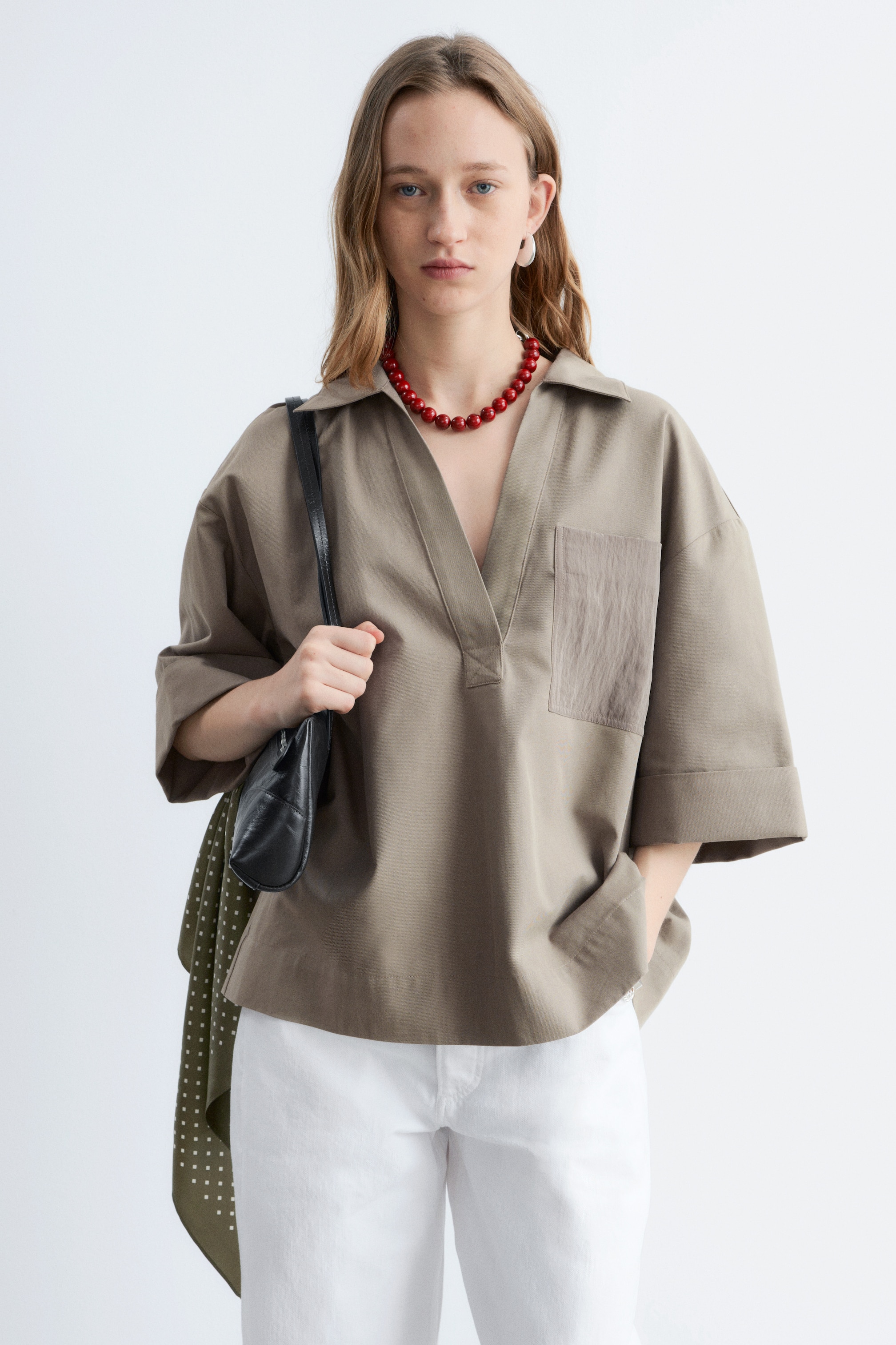 Zobrazit větší obrázek: Khaki popover košile s výrazným límečkem a otevřeným V-čkovým zapínáním má široké, ohrnuté rukávy po lokty. Na pravé straně hrudi je viditelná jediná texturovaná náprsní kapsa a košile volně splývá přes bílé kalhoty.