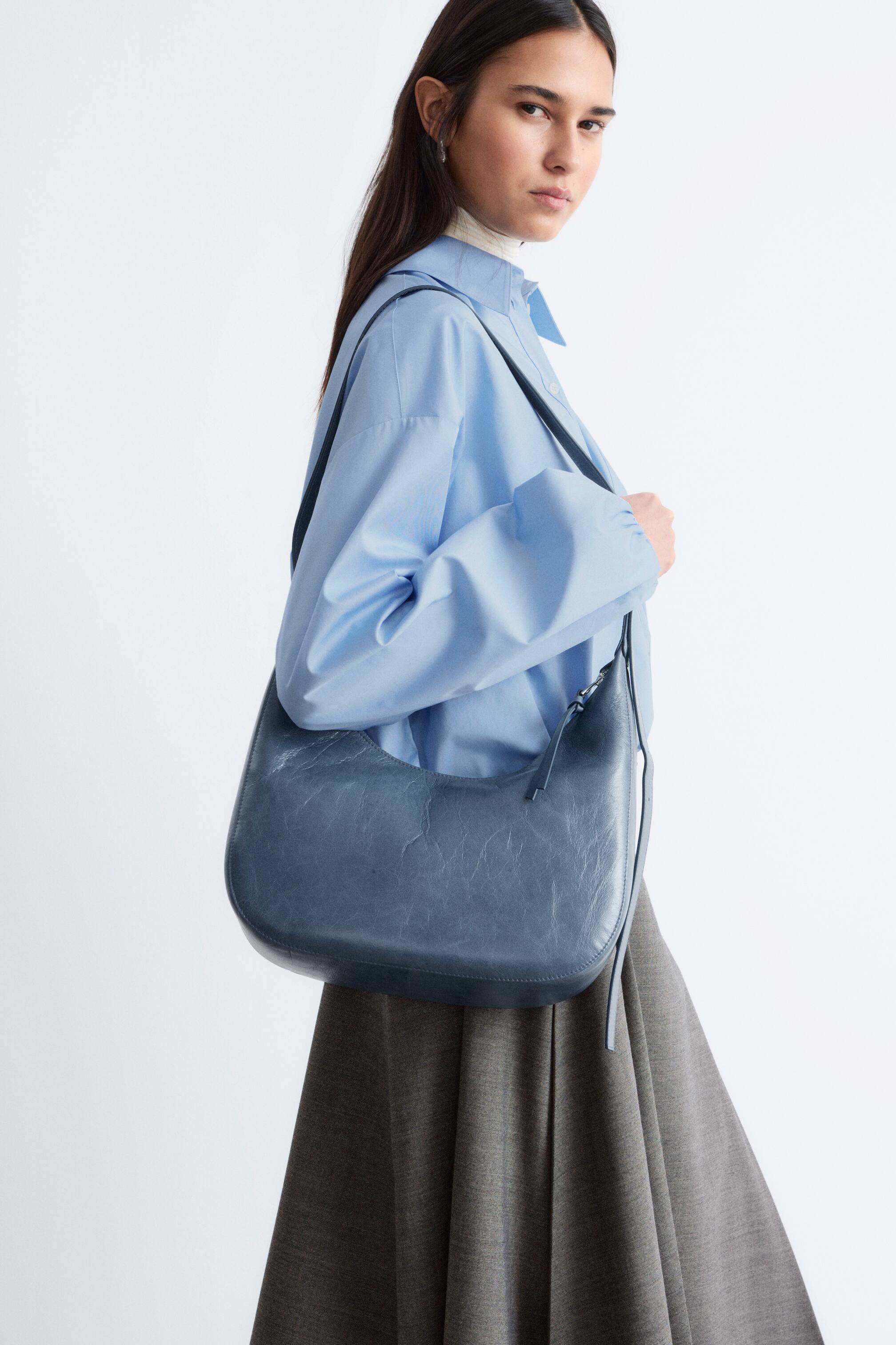 Ver imagen más grande: Un bolso de hombro en forma de media luna de color azul pizarra, con un acabado texturizado y ligeramente arrugado, se lleva sobre el hombro derecho y descansa contra la cadera.