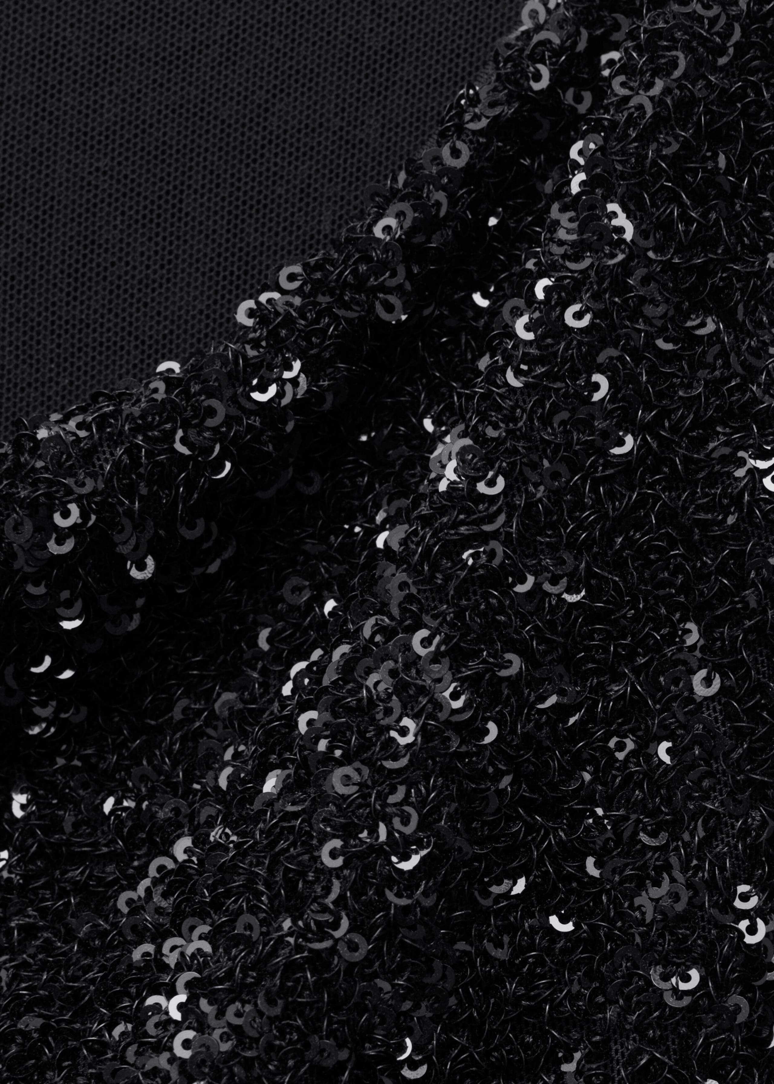A close-up of black fabric showing densely embroidered, shiny black sequins on a sheer black mesh backing visible in the upper-left corner. The fabric drapes slightly to highlight the textured, reflective surface of the sequins.