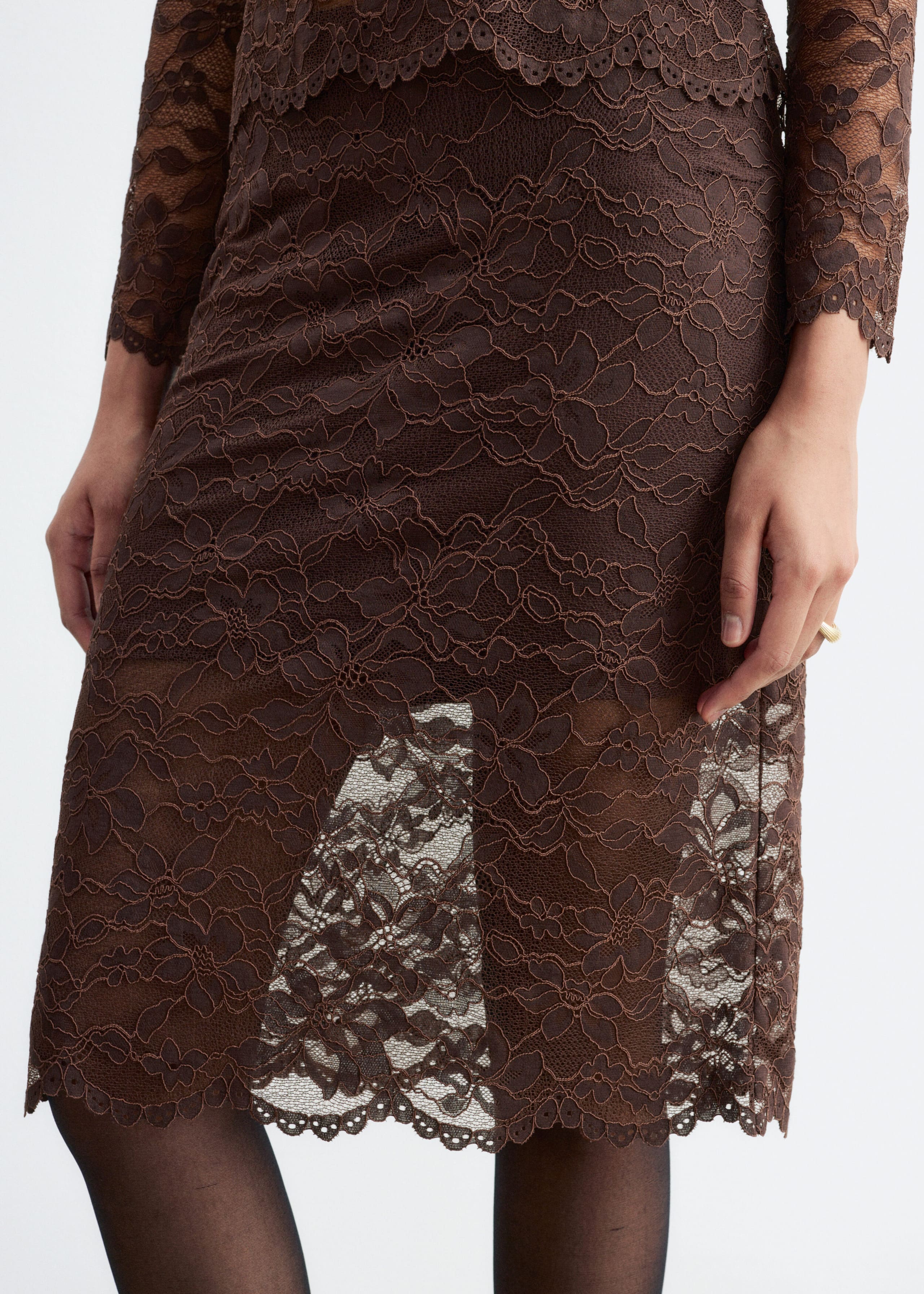 A brown lace skirt with a floral pattern and a scalloped hem is being worn by the model. The knee-length skirt features a sheer lower section over a shorter inner lining and is paired with a matching long-sleeved lace top and sheer black tights.