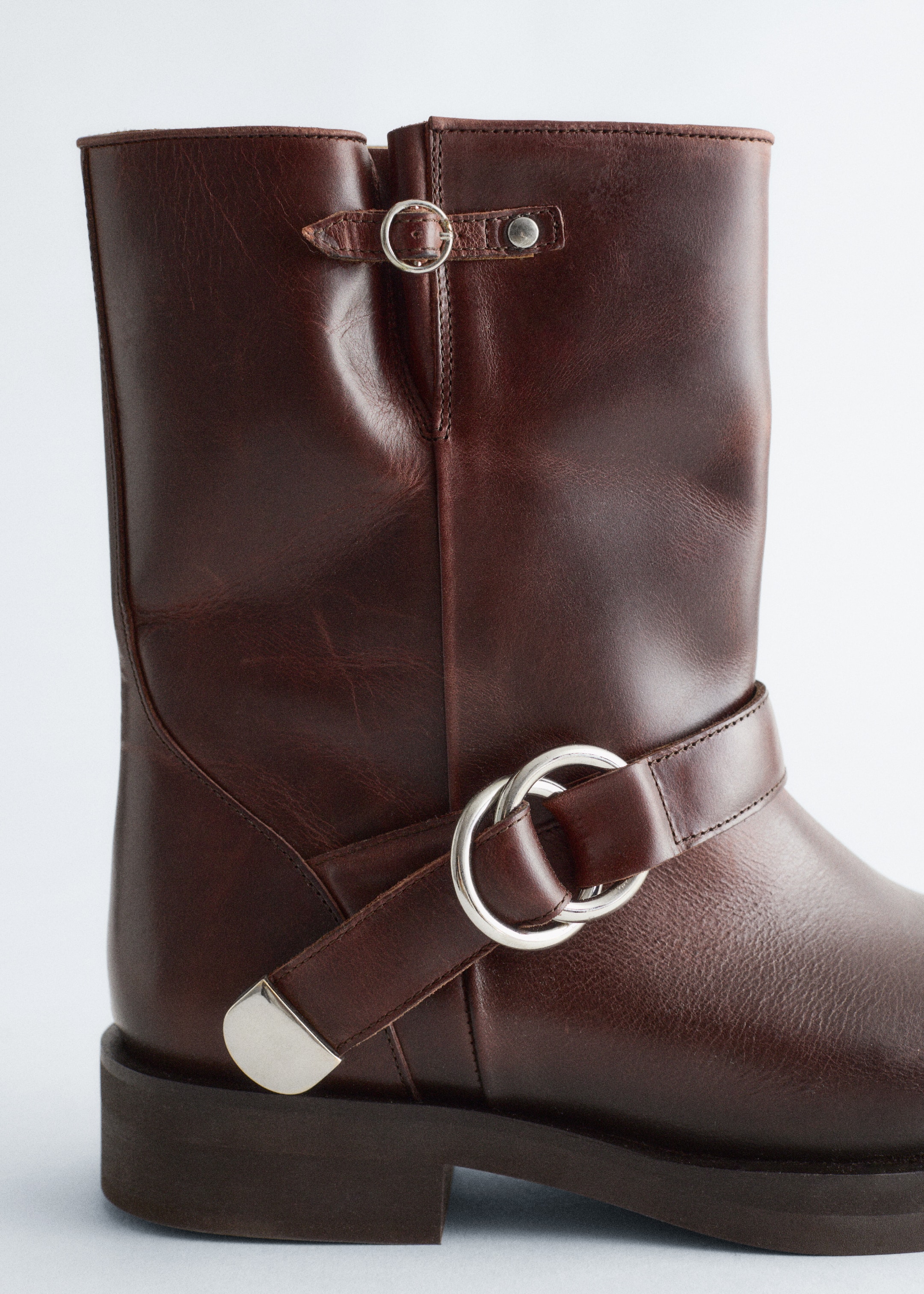 Größeres Bild ansehen: Ein dunkelbrauner Lederstiefel wird aus der Seitenansicht präsentiert, mit einem mittelhohen Schaft und einer klobigen Sohle. Er ist mit silberfarbenen Metallbeschlägen verziert, darunter ein Schnallenriemen nahe der oberen Öffnung und ein dekorativer Riemen mit zwei ineinandergreifenden Ringen um den Knöchel, abgeschlossen mit einer Metallkappe auf der Außenseite.