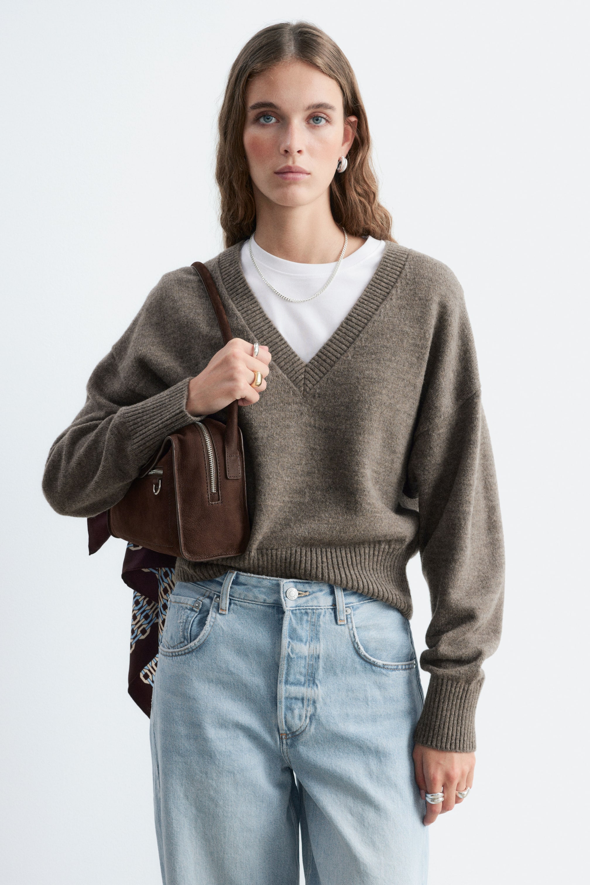 Größeres Bild ansehen: Ein V-Ausschnitt-Pullover aus Wolle und Baumwolle in strukturierter brauner Strickoptik wird vom Model über einem weißen T-Shirt mit Rundhalsausschnitt getragen. Der Pullover verfügt über einen gerippten V-Ausschnitt, überschnittene Schultern sowie gerippte Abschlüsse an den Ärmelbündchen und am Saum und ist mit hell gewaschenen Jeans und einer braunen Umhängetasche kombiniert.