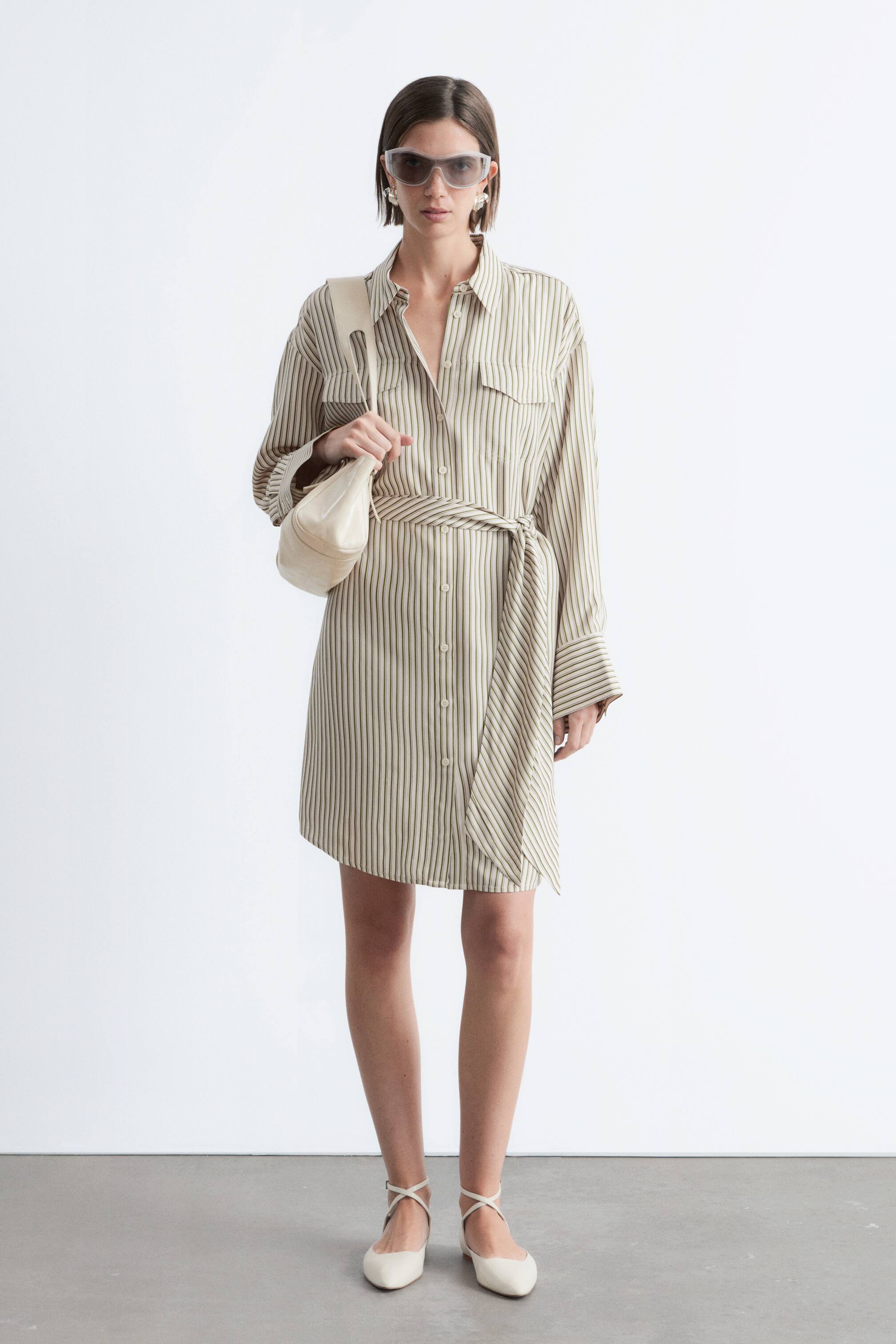 Agrandir l'image: Une robe-chemise à rayures verticales de couleur claire est dotée d'un col en pointe, d'une fermeture boutonnée sur le devant avec deux poches poitrine à rabat, et de manches longues avec des poignets larges. La robe se resserre à la taille avec une ceinture à nouer et présente un ourlet asymétrique.