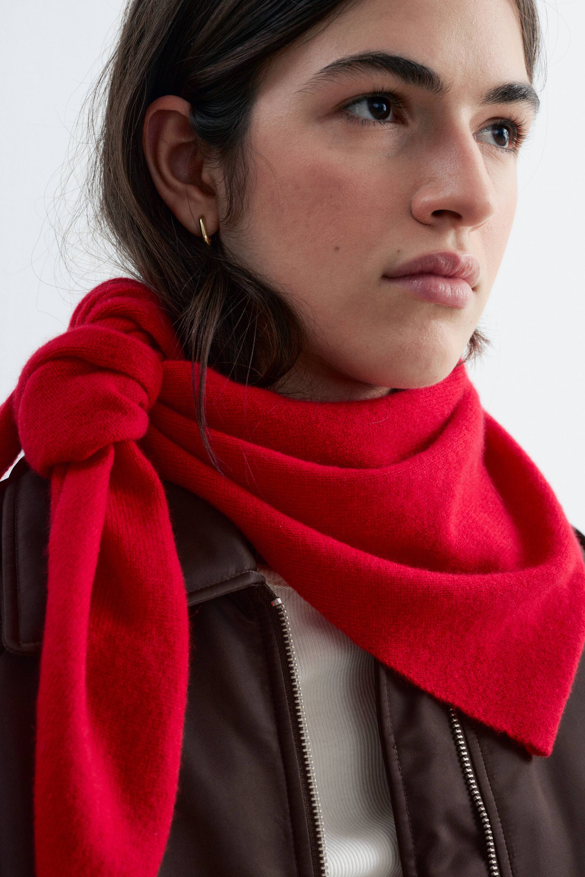 Agrandir l'image: Un foulard triangulaire en maille rouge est porté par le mannequin, drapé autour du cou avec la pointe du triangle orientée vers l’avant et les extrémités nouées de façon lâche sur un côté. Le foulard est porté par-dessus une veste marron foncé zippée.