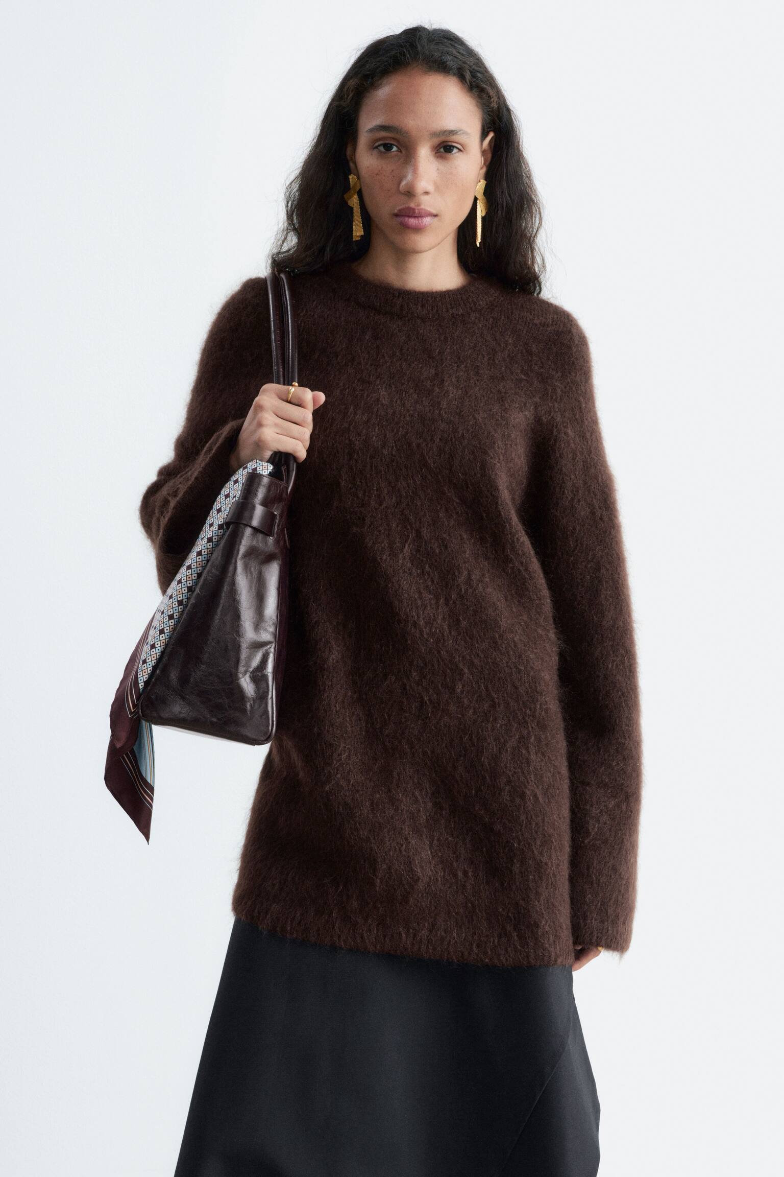 Un pull col rond en maille duveteuse marron foncÃ© est portÃ© par le mannequin. Le mannequin porte un sac bandouliÃ¨re en cuir marron foncÃ© avec une Ã©charpe Ã  motif nouÃ©e Ã  la bandouliÃ¨re, portÃ© avec une jupe noire. - 1