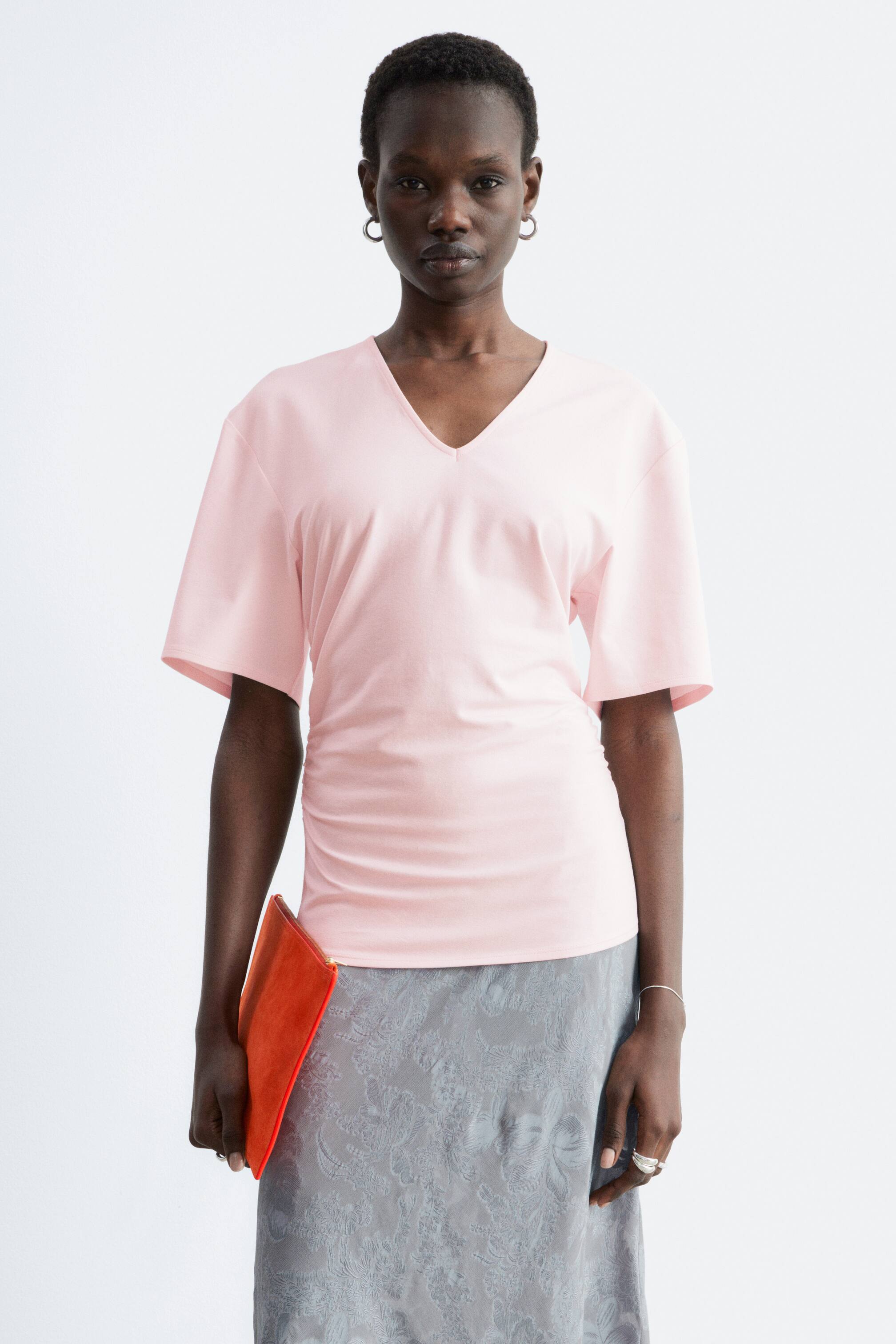 Προβολή μεγαλύτερης εικόνας: Ένα ανοιχτό ροζ τοπ με V λαιμόκοψη, διαθέτει V λαιμόκοψη, κοντά φαρδιά μανίκια και λεπτομέρειες με πιέτες κατά μήκος της αριστερής πλευράς, προσφέροντας μια χαλαρή εφαρμογή. Το στρίφωμα του τοπ πέφτει μέχρι το ισχίο, αγκαλιάζοντας μια φούστα με γκρι μοτίβο.