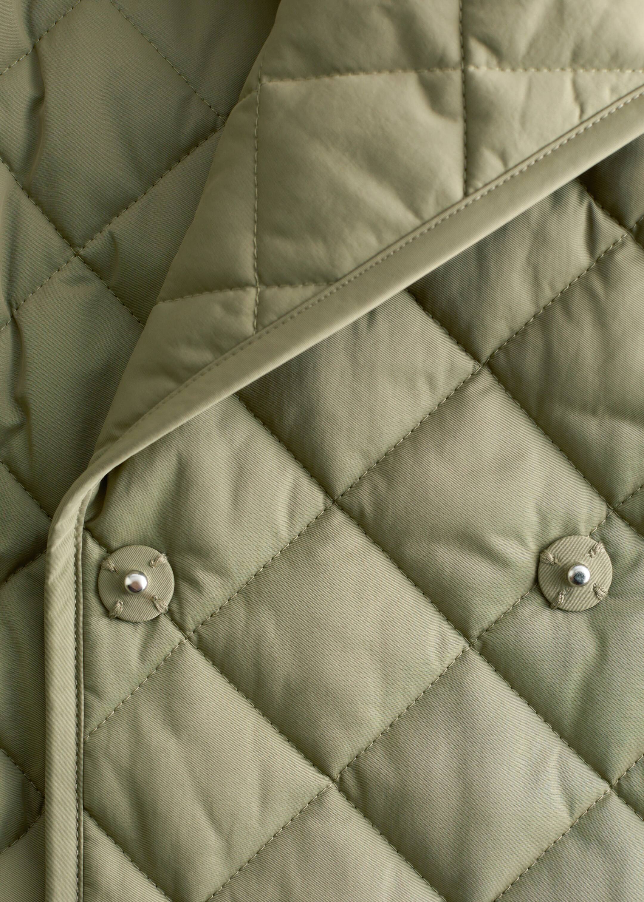 View larger image: An olive green quilted jacket is featuring a diamond pattern with light padding. Two silver-toned press buttons are visibly securing a smooth, unquilted fabric edge that is folding over the jacket's main quilted surface.