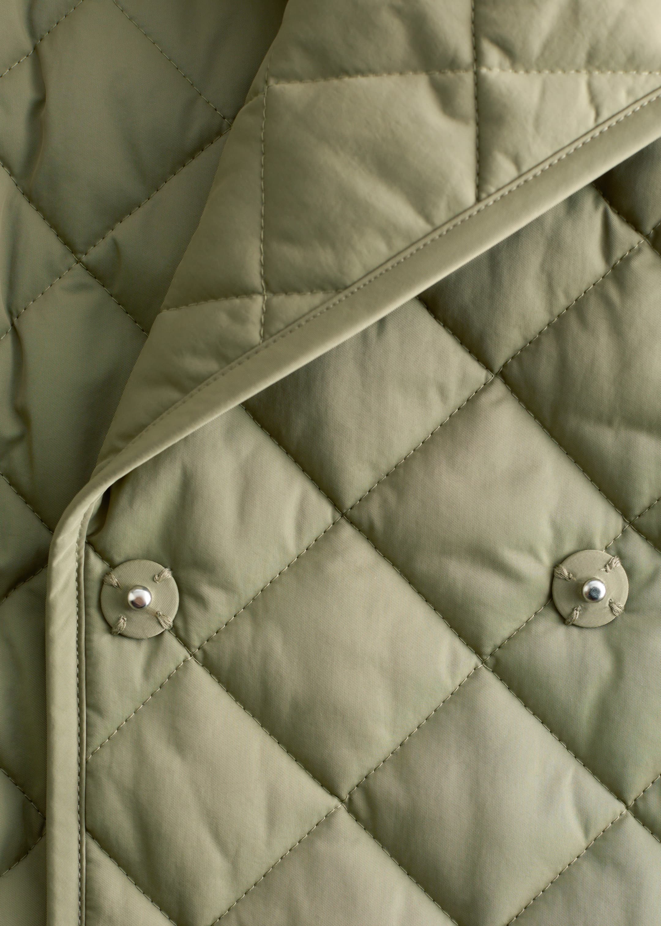 Visualizza immagine più grande: Una giacca trapuntata verde oliva presenta un motivo a diamante con un'imbottitura leggera. Due bottoni a pressione in tonalità argento fissano visibilmente un bordo di tessuto liscio e non trapuntato che si piega sulla superficie principale trapuntata della giacca.