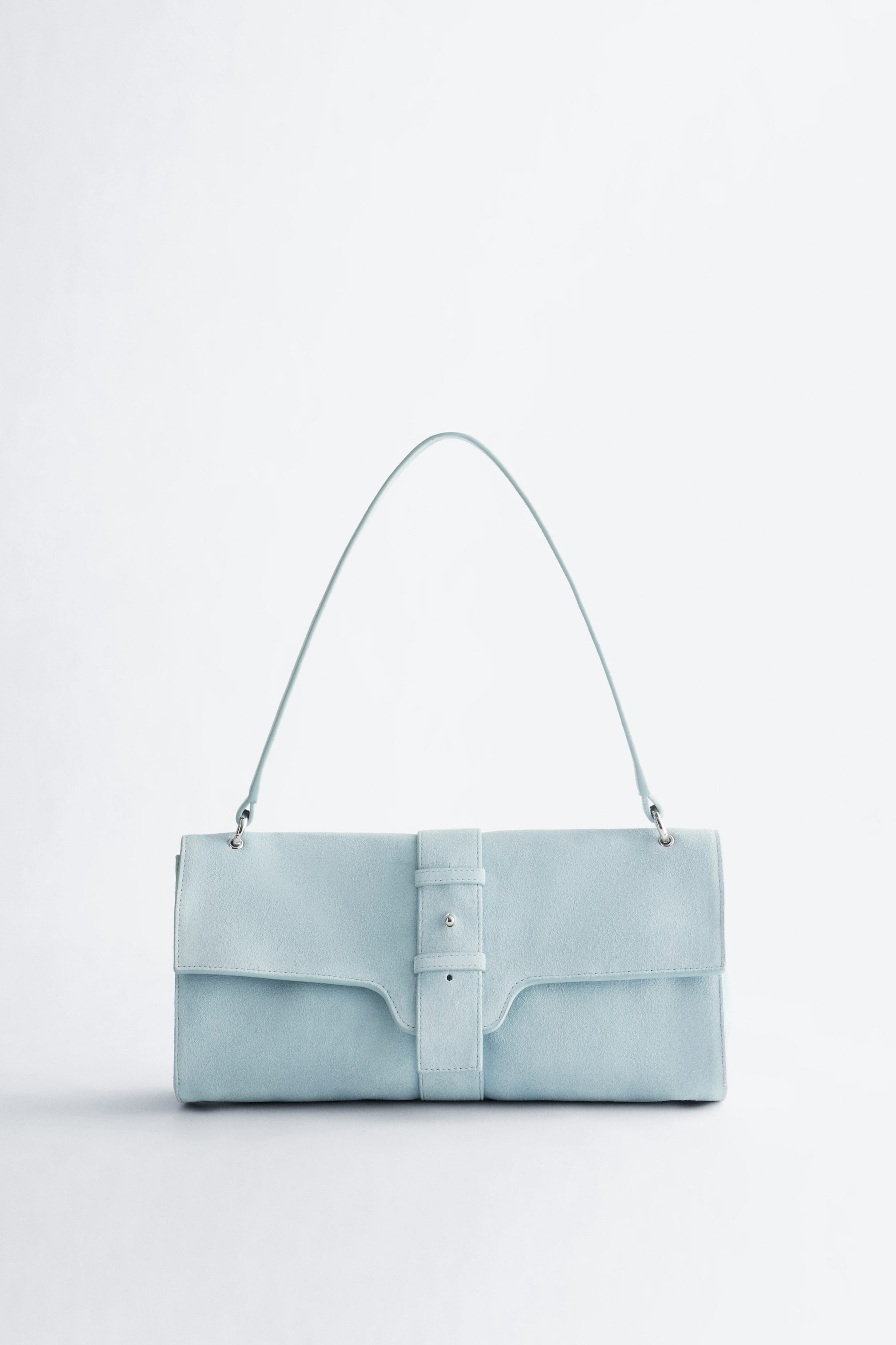 Agrandir l'image: Un sac à bandoulière bleu clair est présenté de face, avec un corps rectangulaire allongé et une texture semblable à du daim. Un rabat profilé est sécurisé par une sangle verticale centrale avec un bouton-pression argenté, et une fine bandoulière assortie s'arque au-dessus du sac.