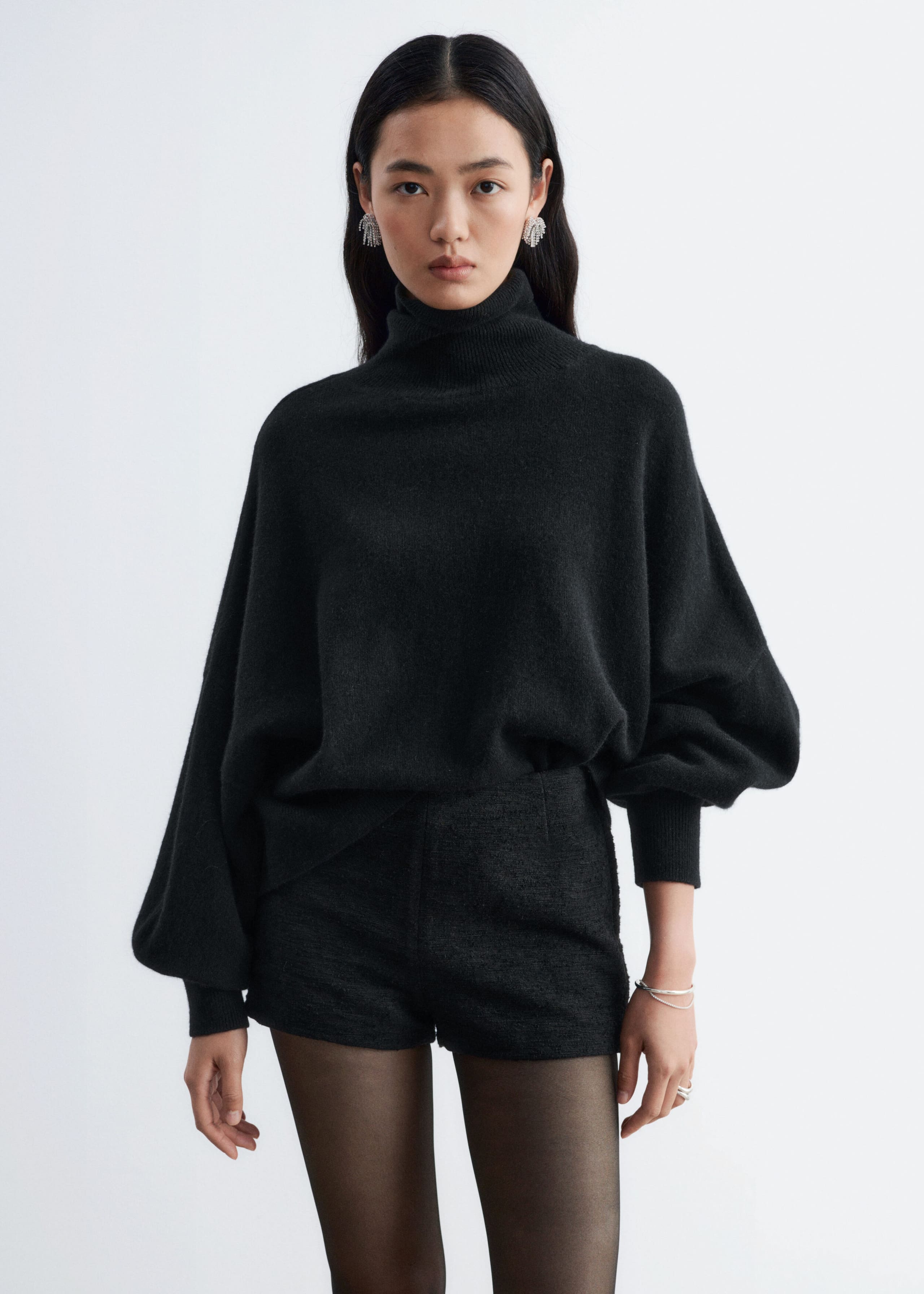These black tweed shorts are worn by the model and styled with a loose-fitting black roll-neck jumper tucked into the front waistband, alongside sheer black tights. The high-waisted shorts feature a textured fabric and structured darts at the waist for a defined silhouette.