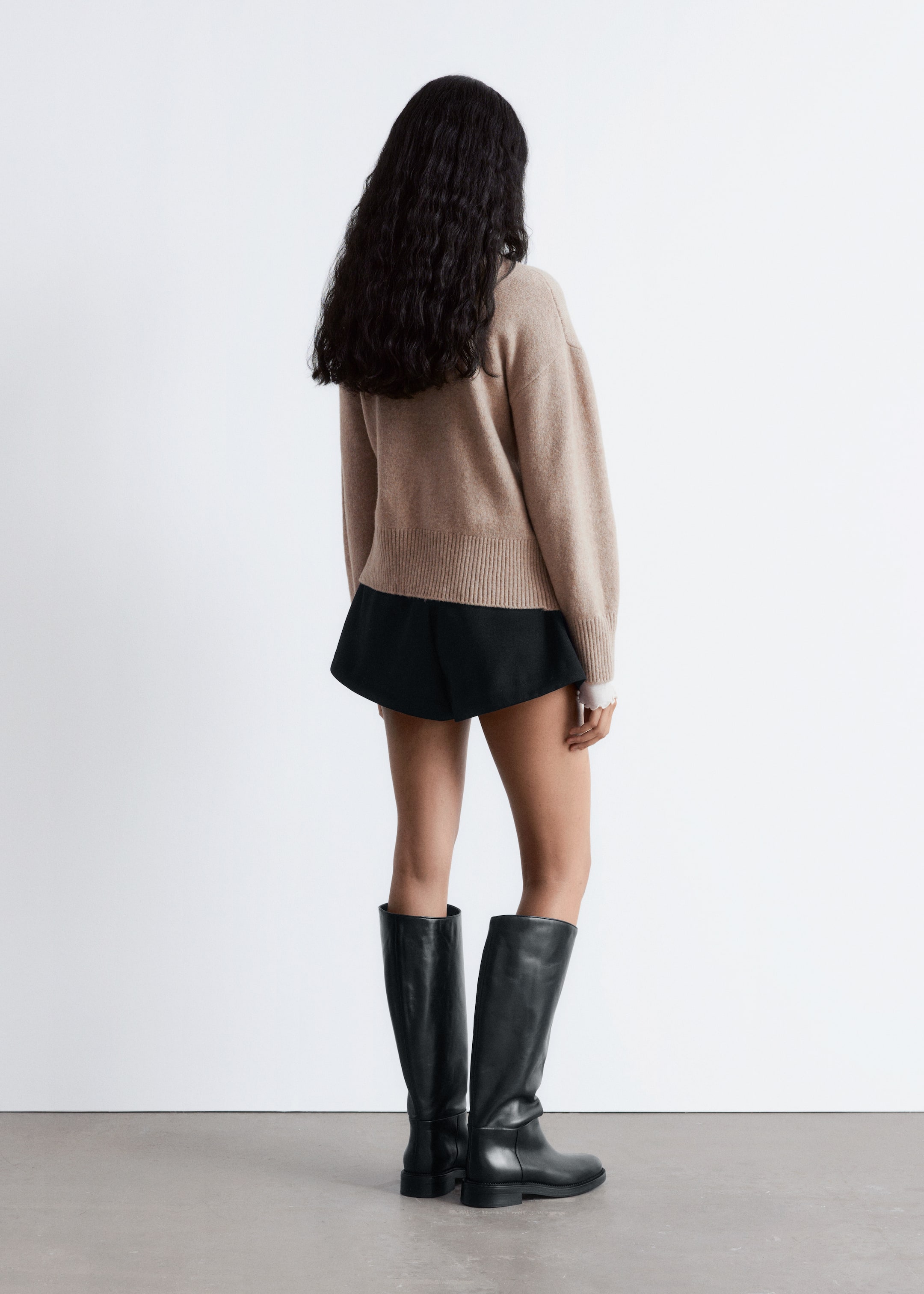 Größeres Bild anzeigen: Ein hellbrauner, gerippter Strickpullover wird getragen, von hinten betrachtet, mit einem weißen Bündchen, das subtil aus seinem langen Ärmel hervorschaut. Der Pullover wird mit kurzen schwarzen ausgestellten Shorts und hohen schwarzen Lederstiefeln kombiniert.