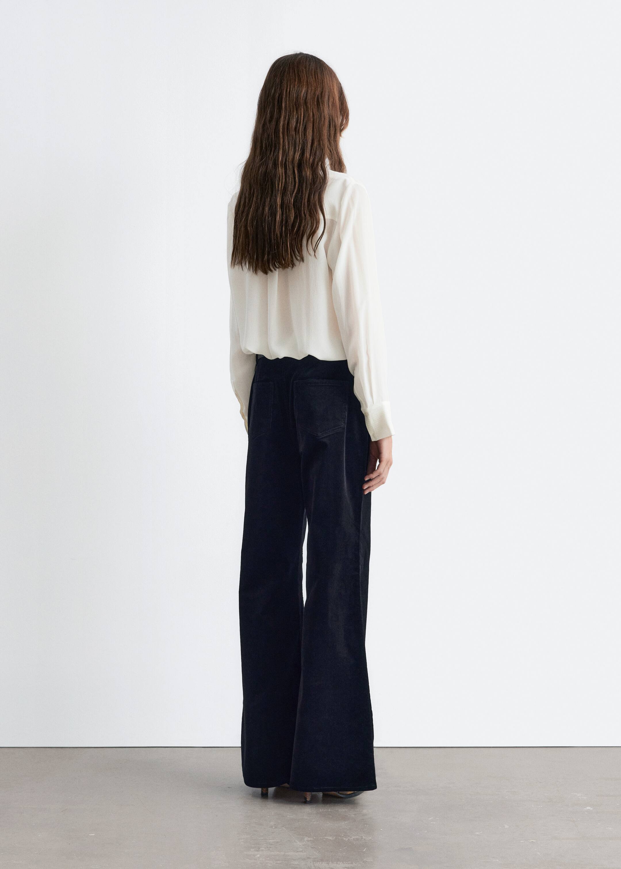 Grotere afbeelding bekijken: Donkere, wijde corduroy broek wordt van achteren getoond, met twee zichtbare opgestikte zakken. Ze vloeien naar de vloer en worden gedragen met een lichtgekleurde blouse met lange mouwen die in de tailleband is gestopt.