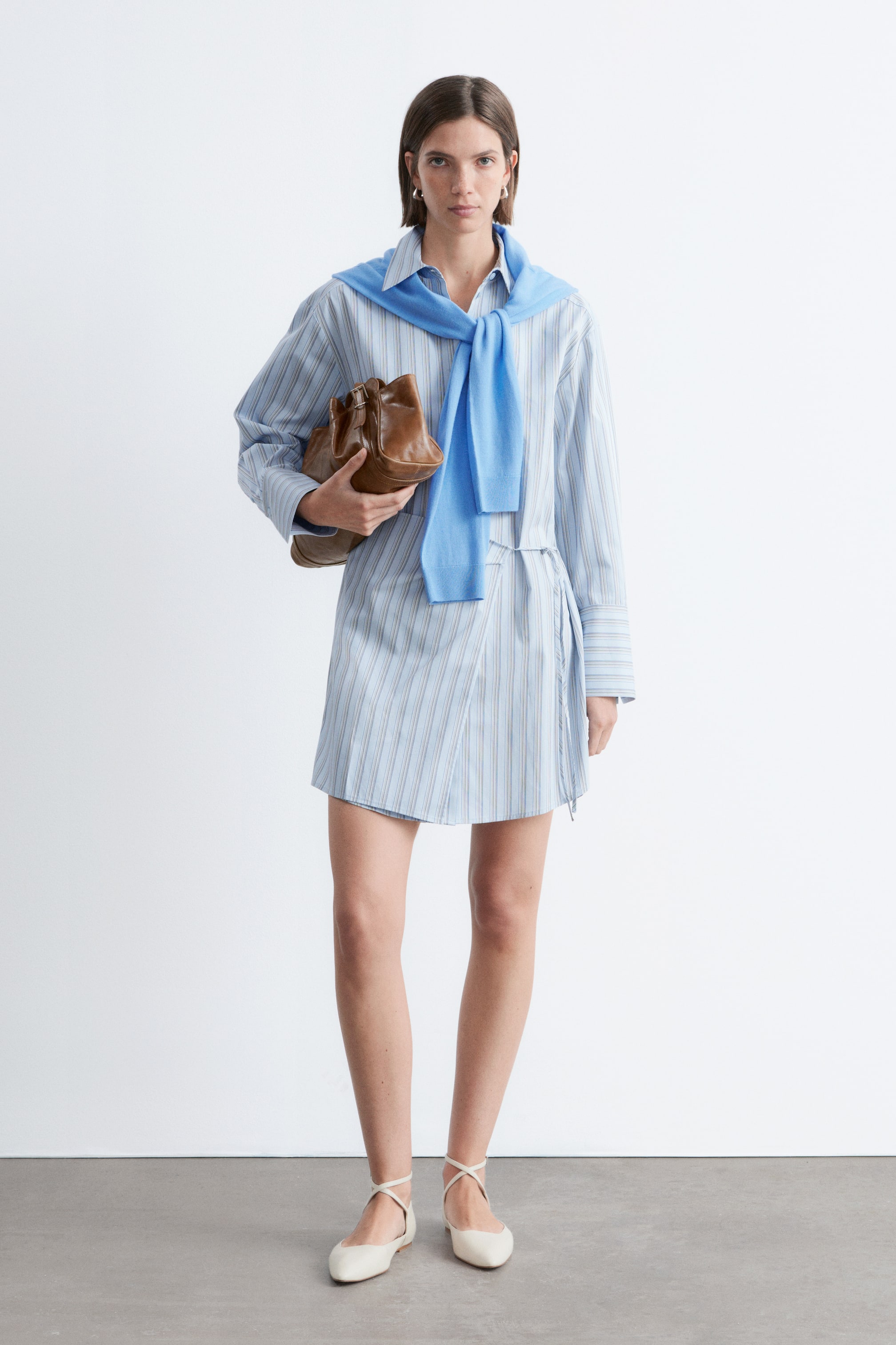 Visualizza immagine più grande: Un abito camicia a portafoglio azzurro con sottili righe verticali bianche e beige è presentato frontalmente, caratterizzato da un colletto e maniche lunghe con polsini abbottonati. Un maglione blu è drappeggiato sulle spalle, parzialmente coprendo la parte superiore, e l'abito si annoda in vita, creando una gonna sovrapposta.